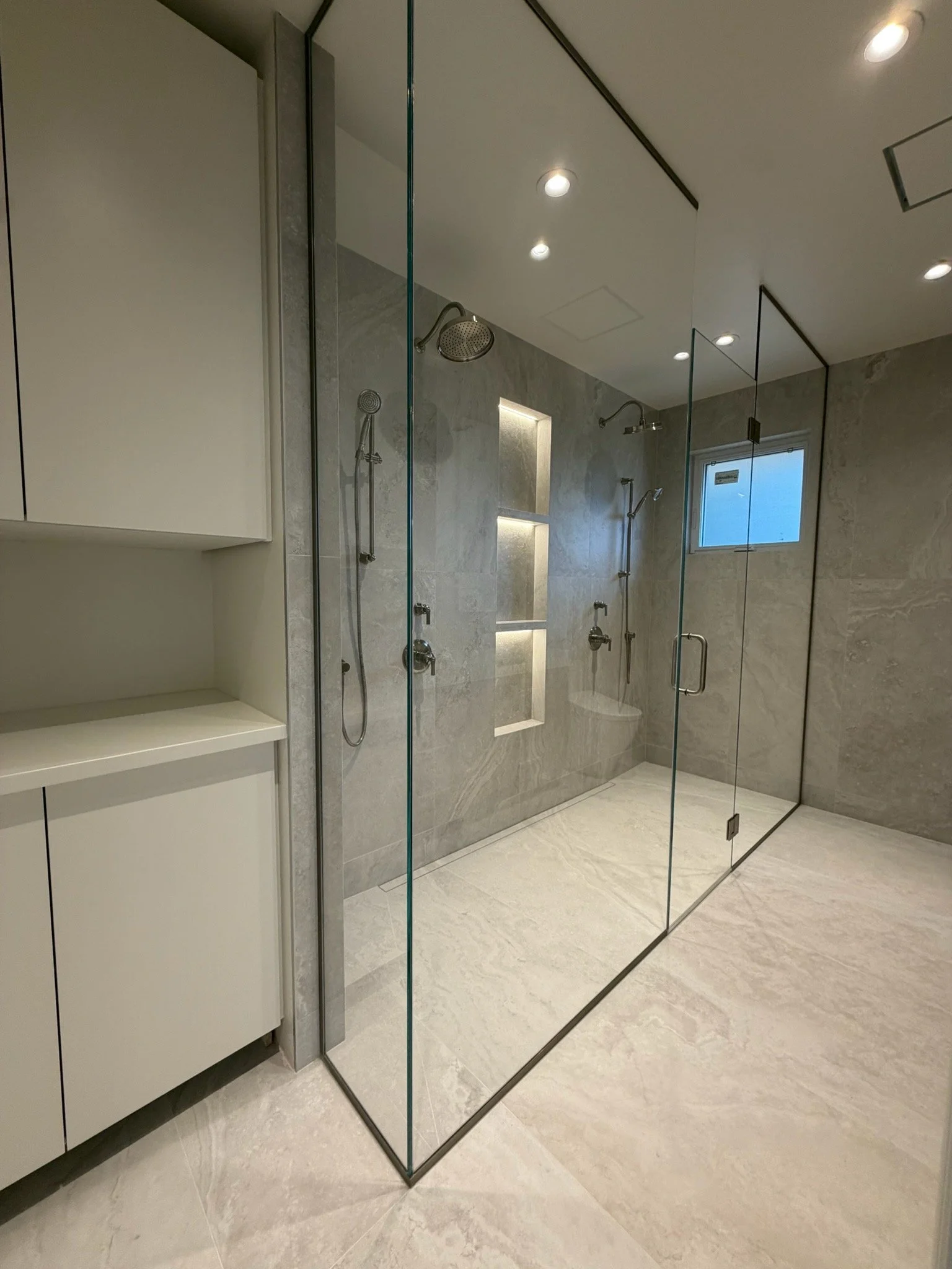 Modern bathroom with a glass-enclosed shower, gray marble walls, and ceiling-mounted showerheads. There is a window and built-in shelves with lighting inside the shower.