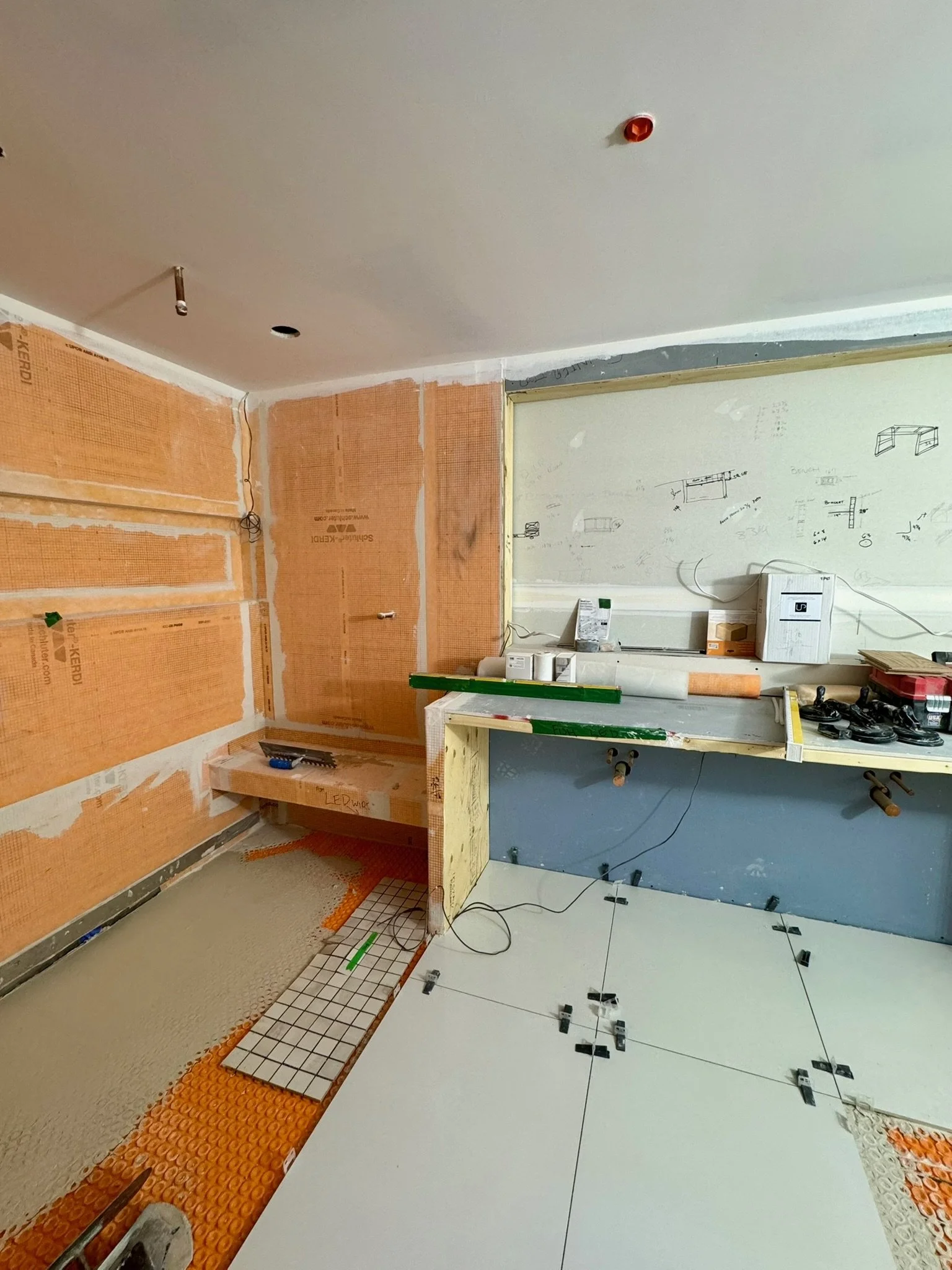 Interior construction site with partially tiled floor, drywall walls, and exposed ceiling, equipment, tools, and materials scattered around.