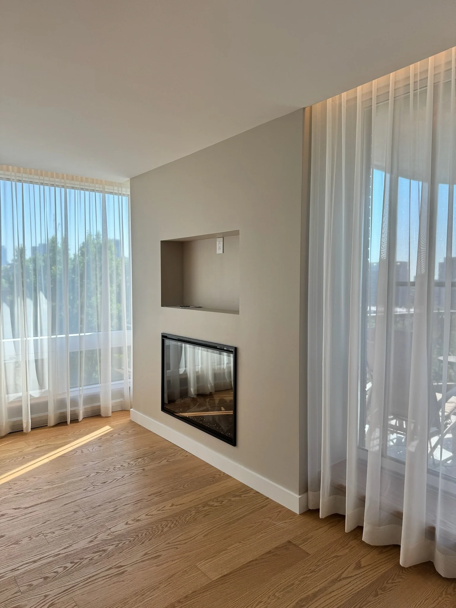 A sunlit modern living room corner with light wood flooring, large windows with sheer white curtains, and a wall-mounted electric fireplace with a mirror beneath it.