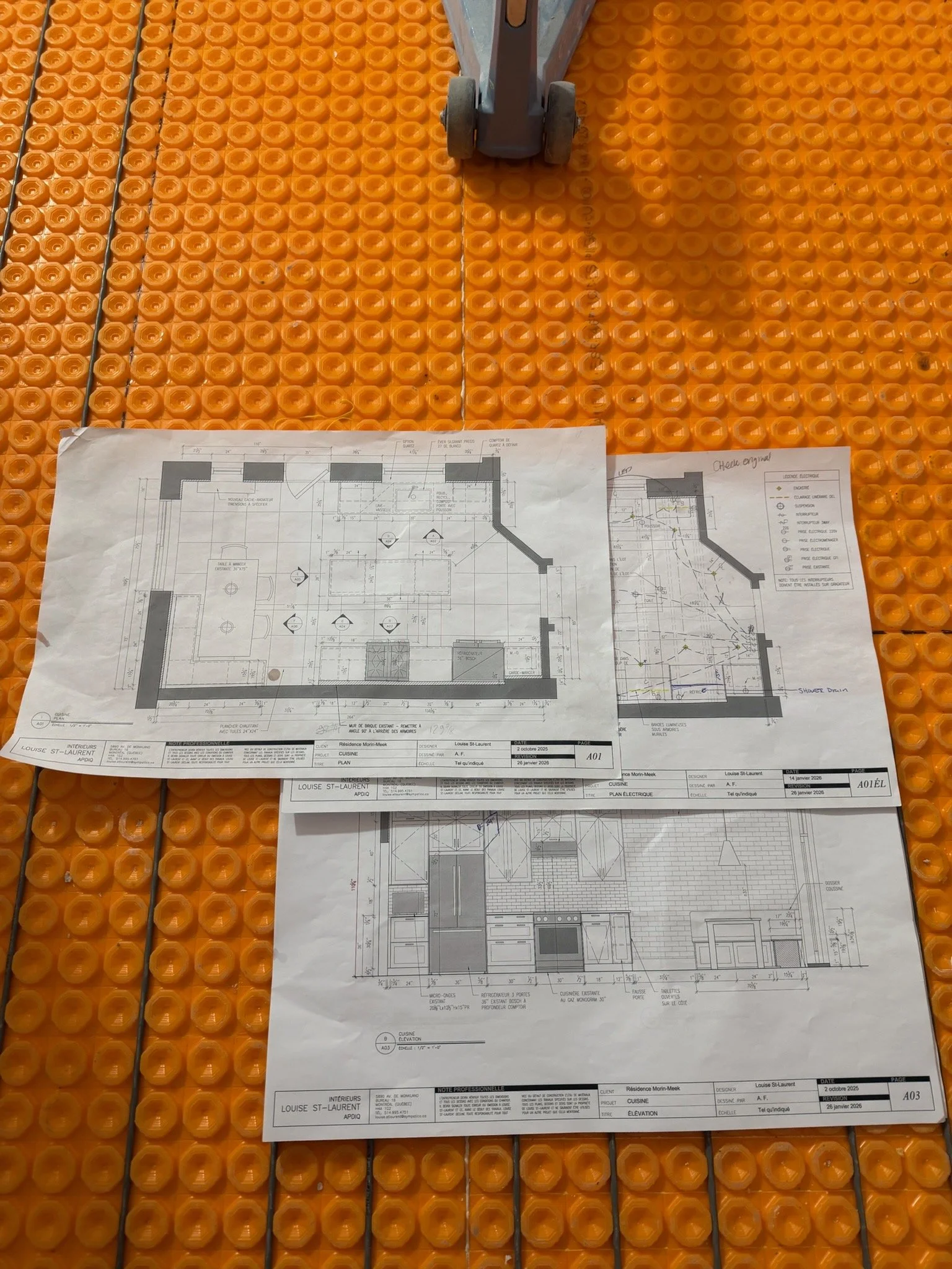 Blueprint plans laid on an orange plastic grid surface with a small caster wheel attached to a metal frame at the top.