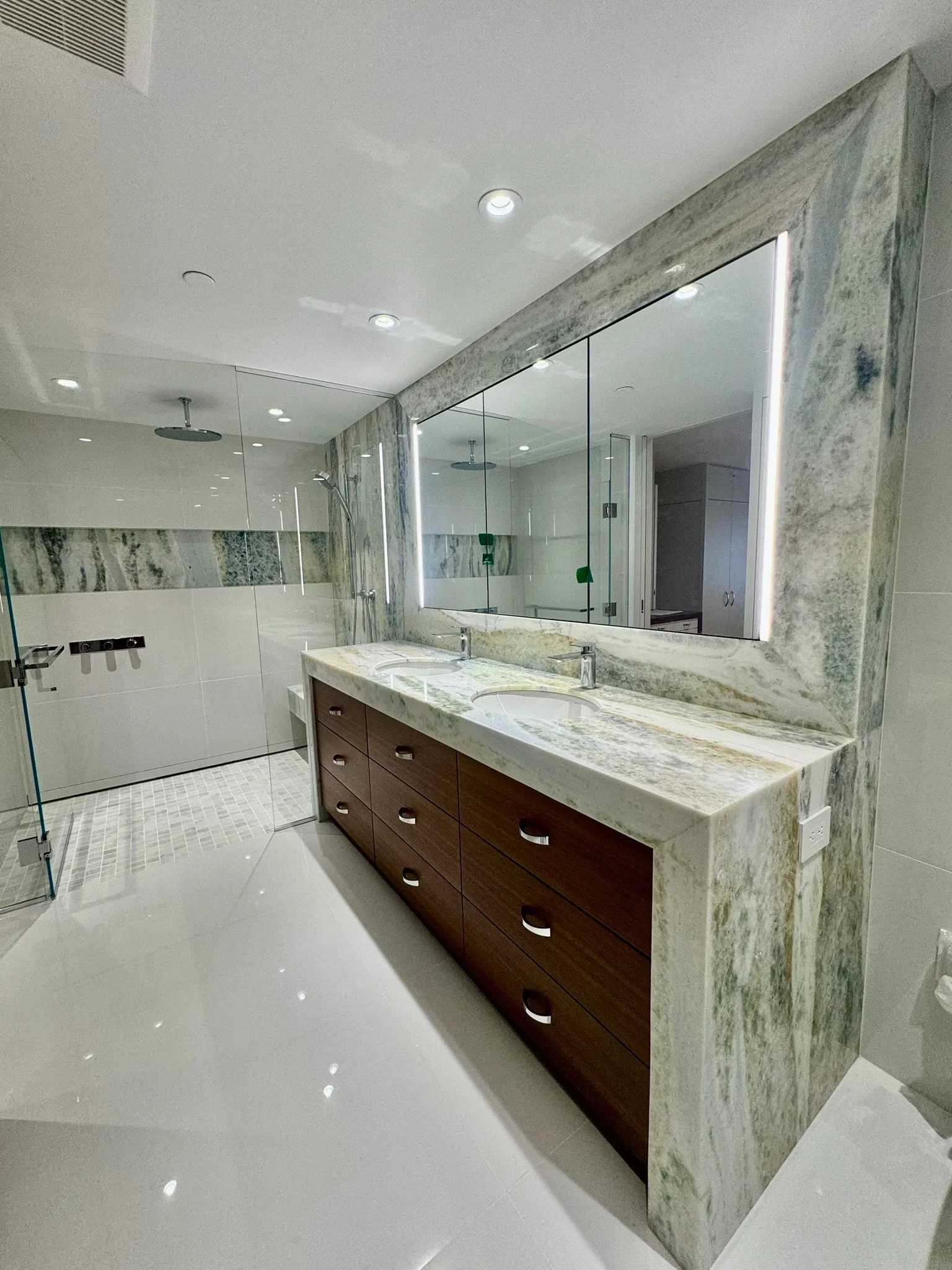 Modern bathroom with a large marble vanity mirror and double sinks. The vanity has a wooden cabinet with multiple drawers. There is a walk-in shower with a glass door and rain showerheads on the wall. The floor is shiny white tile, and recessed light