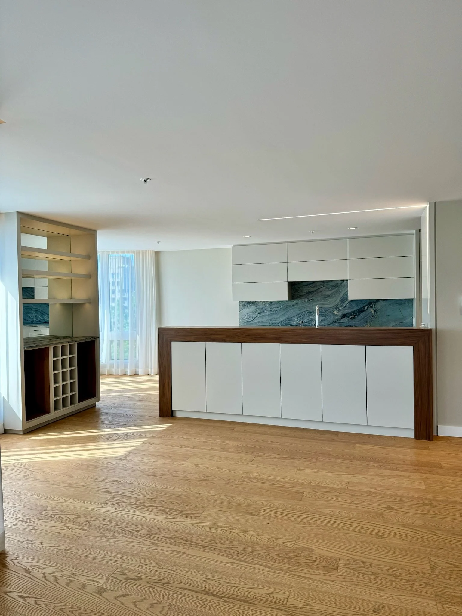 Modern kitchen with light wood flooring, white cabinetry with wood accents, a marble backsplash, and large windows with sheer curtains letting in natural light.