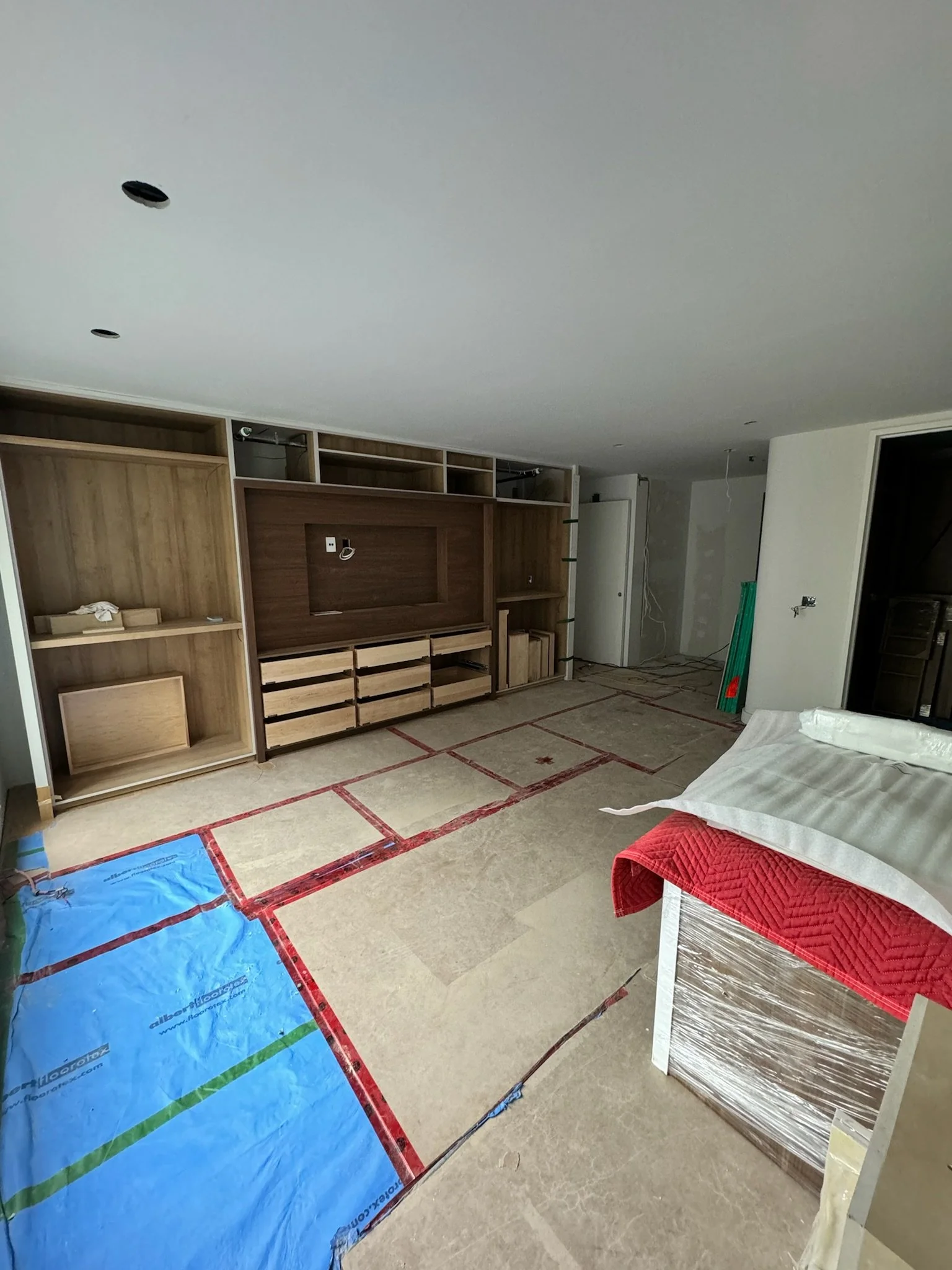 Living room under construction with wooden wall unit, plastic wrapping on furniture, and marked floor for installation