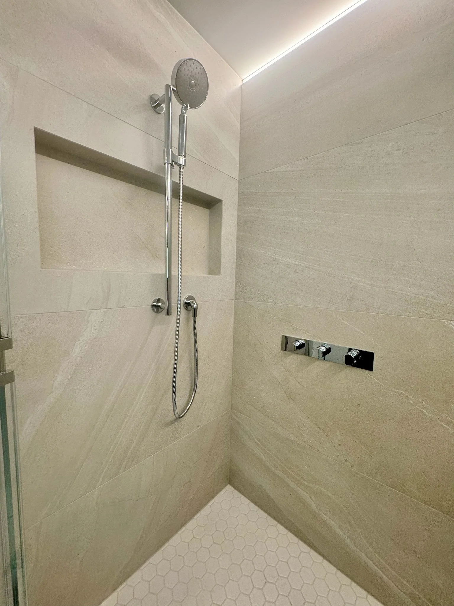 A modern glass-enclosed shower with beige wall tiles, a built-in shower niche, a chrome hand-held showerhead, and a set of chrome controls on the wall.