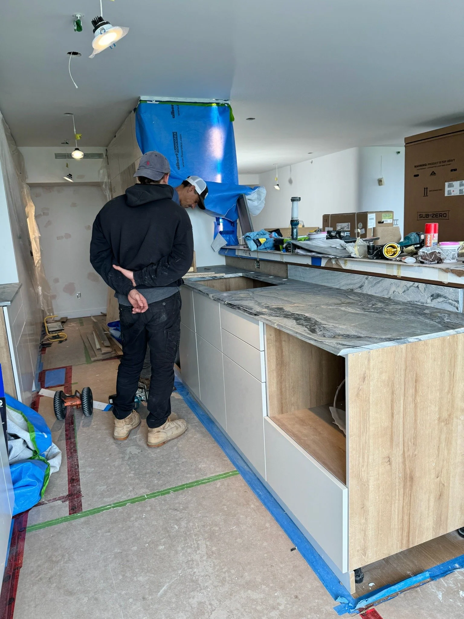 Two workers in a kitchen under construction, installing a marble countertop on white cabinetry with wooden sides. The counter is in a state of installation with tools and materials scattered around.