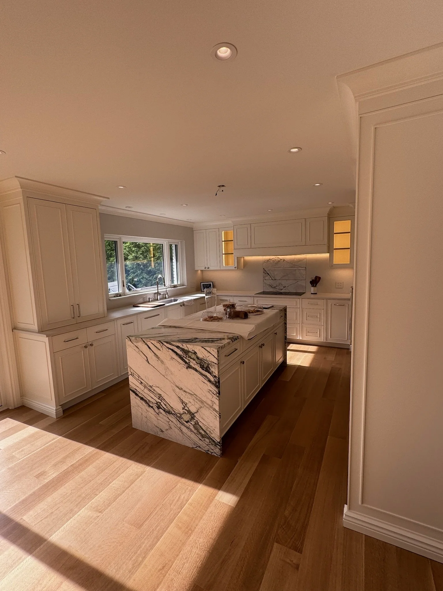A modern kitchen with white cabinets, a marble island, hardwood floors, and a window letting in natural light.