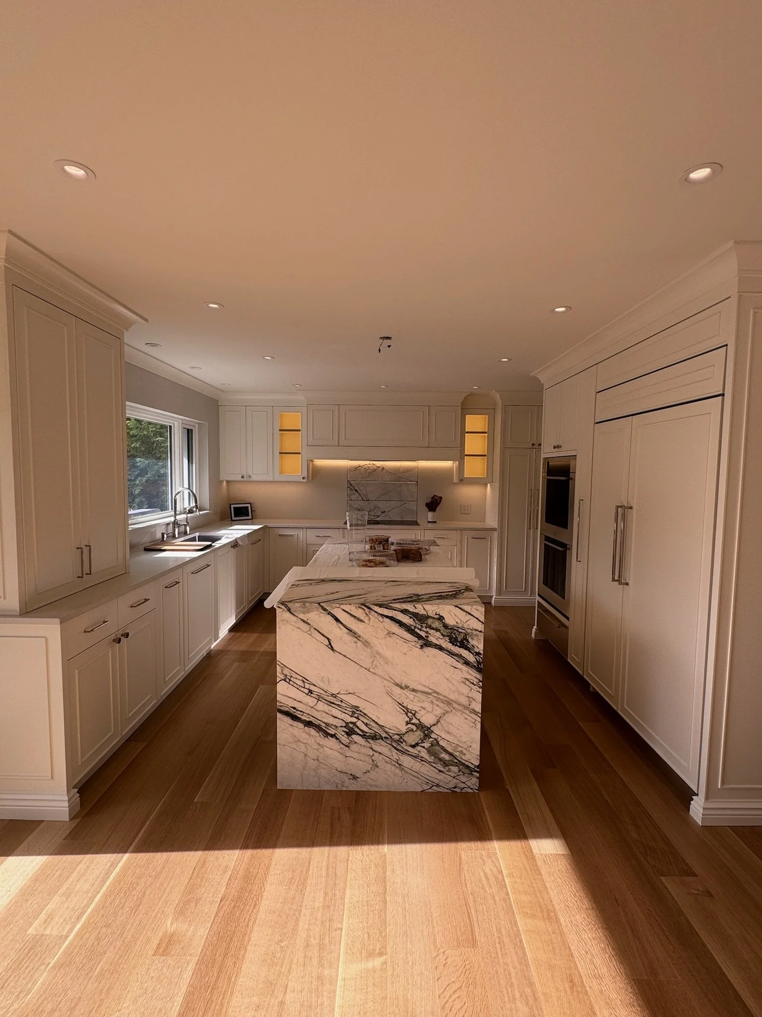 Modern kitchen with off-white cabinetry, marble island, wood flooring, and a large window over the sink.