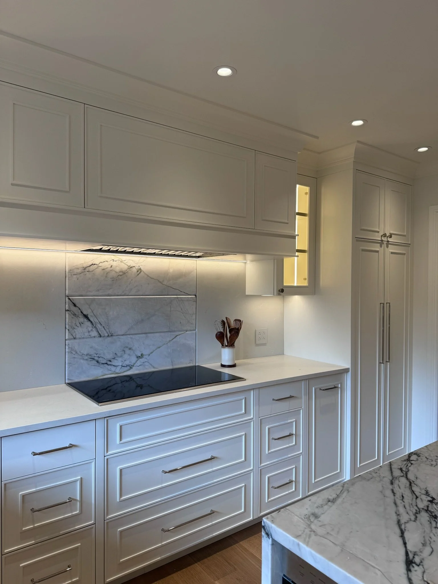 Modern kitchen with white cabinets, marble backsplash, and a black stovetop. The countertop has a utensil holder with wooden spoons and a marble island in the foreground.