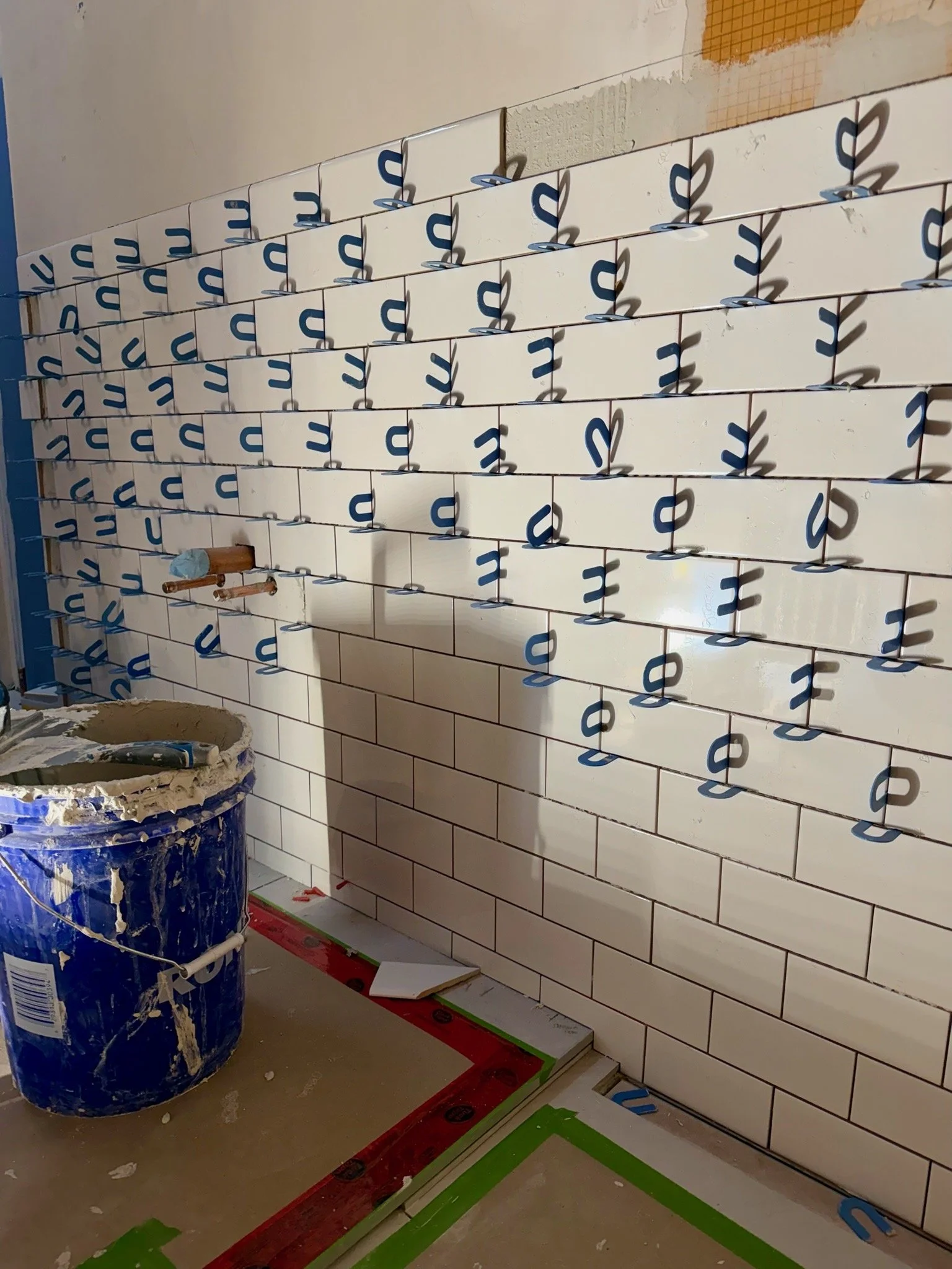 Interior view of a wall under construction with white tiles and blue protective stickers, construction tools, and supplies visible on the floor.