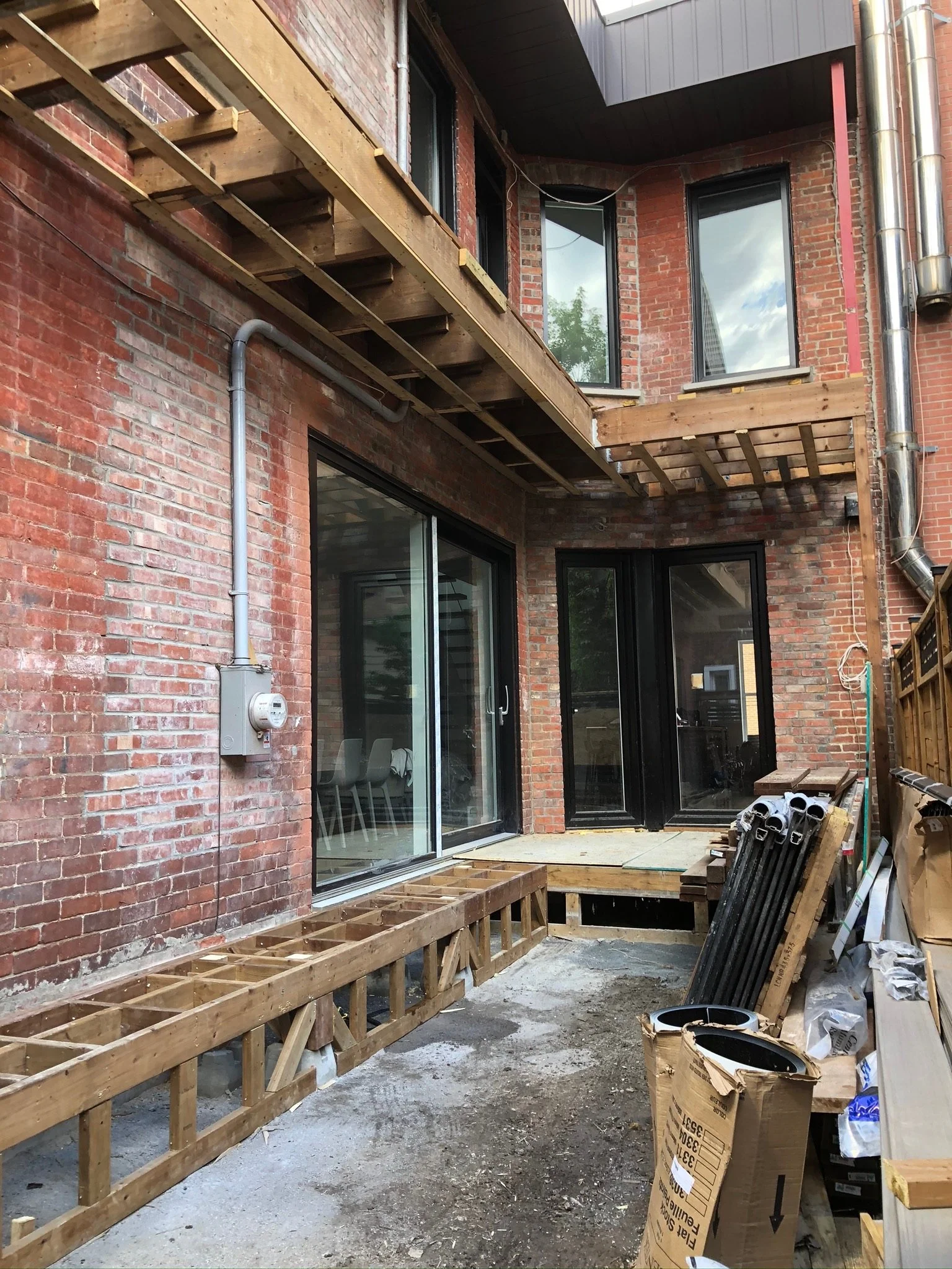 Construction site with wooden framing for deck or porch, brick exterior building, sliding glass door, and construction materials including pipes, boxes, and wooden planks.