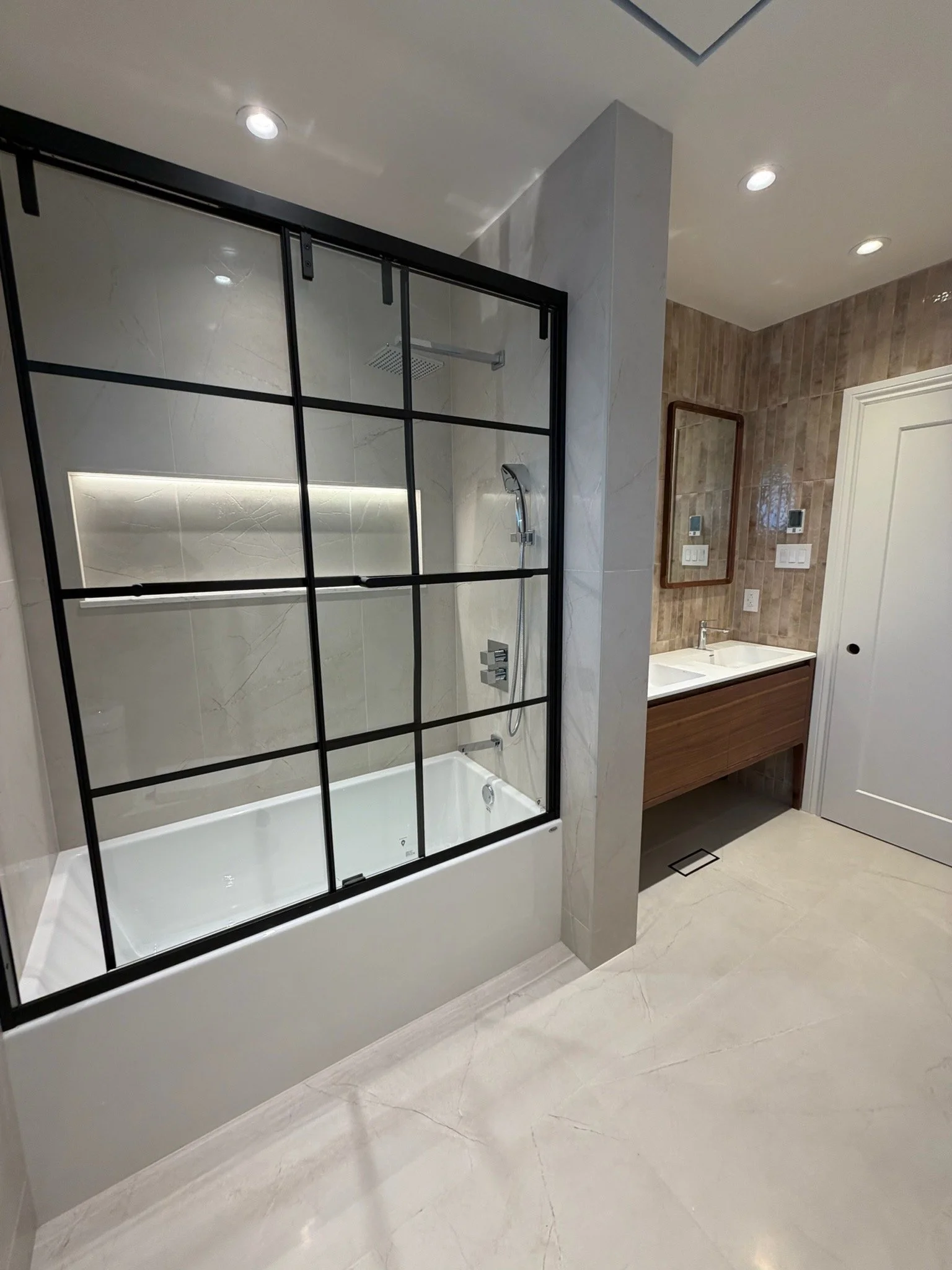 Bathroom with glass shower enclosure, wooden vanity with double sink, wall-mounted mirrors, beige tiled walls, recessed ceiling lights, and beige marble floor.