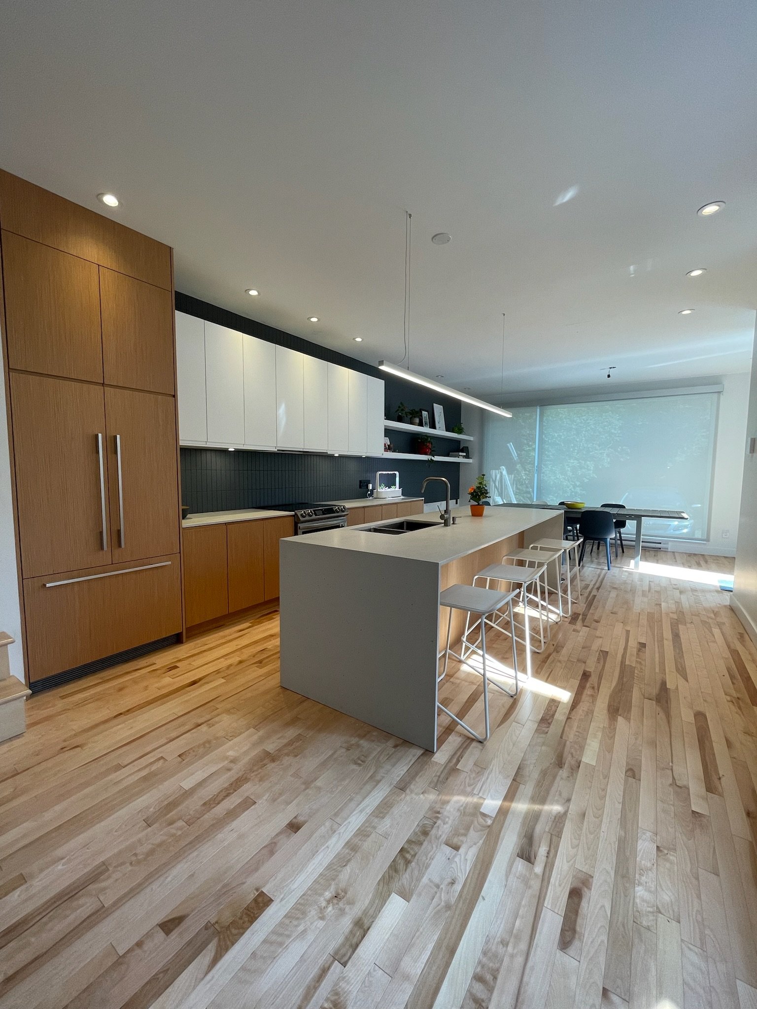 Modern kitchen with wooden flooring, white and wood cabinets, island with bar stools, and large window with a view of greenery.