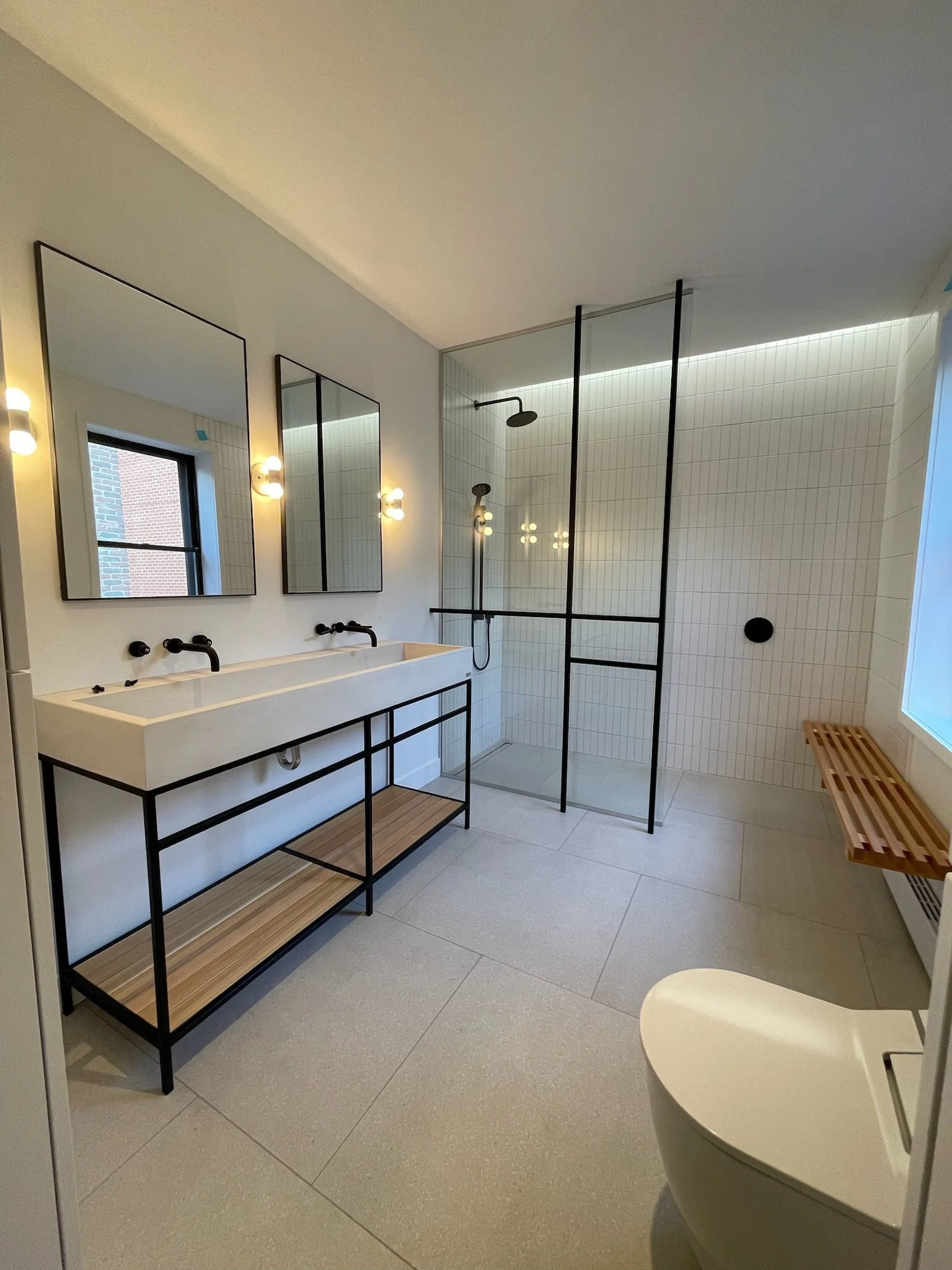 Modern bathroom with white sink and black fixtures, rectangular mirrors, light fixtures, shower with black frame, white tiled walls, wooden bench, and beige tiled floor.
