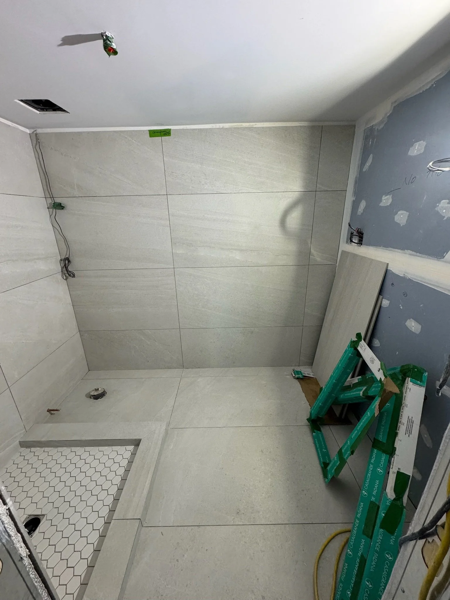 Bathroom under construction with partially tiled floor and wall. Visible tools and materials, including green packaging, wires, and a small section of hexagonal tile floor in the corner.