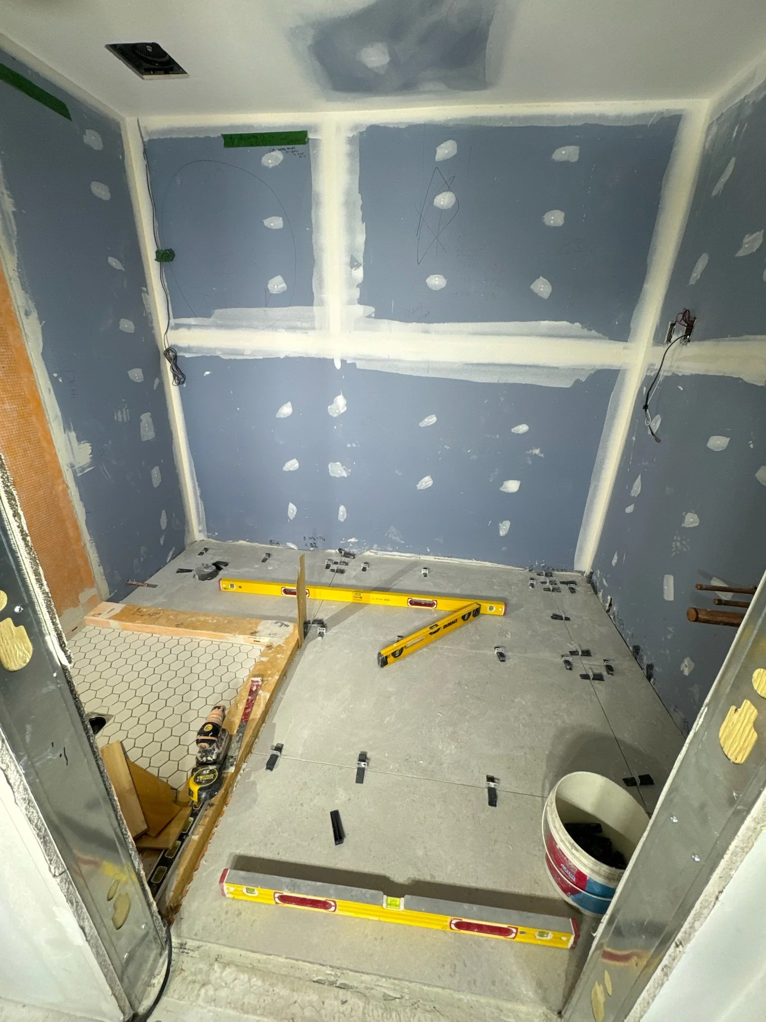 Bathroom under construction with partially installed tile floor, blue drywall with drywall mud, and tools including levels and a bucket
