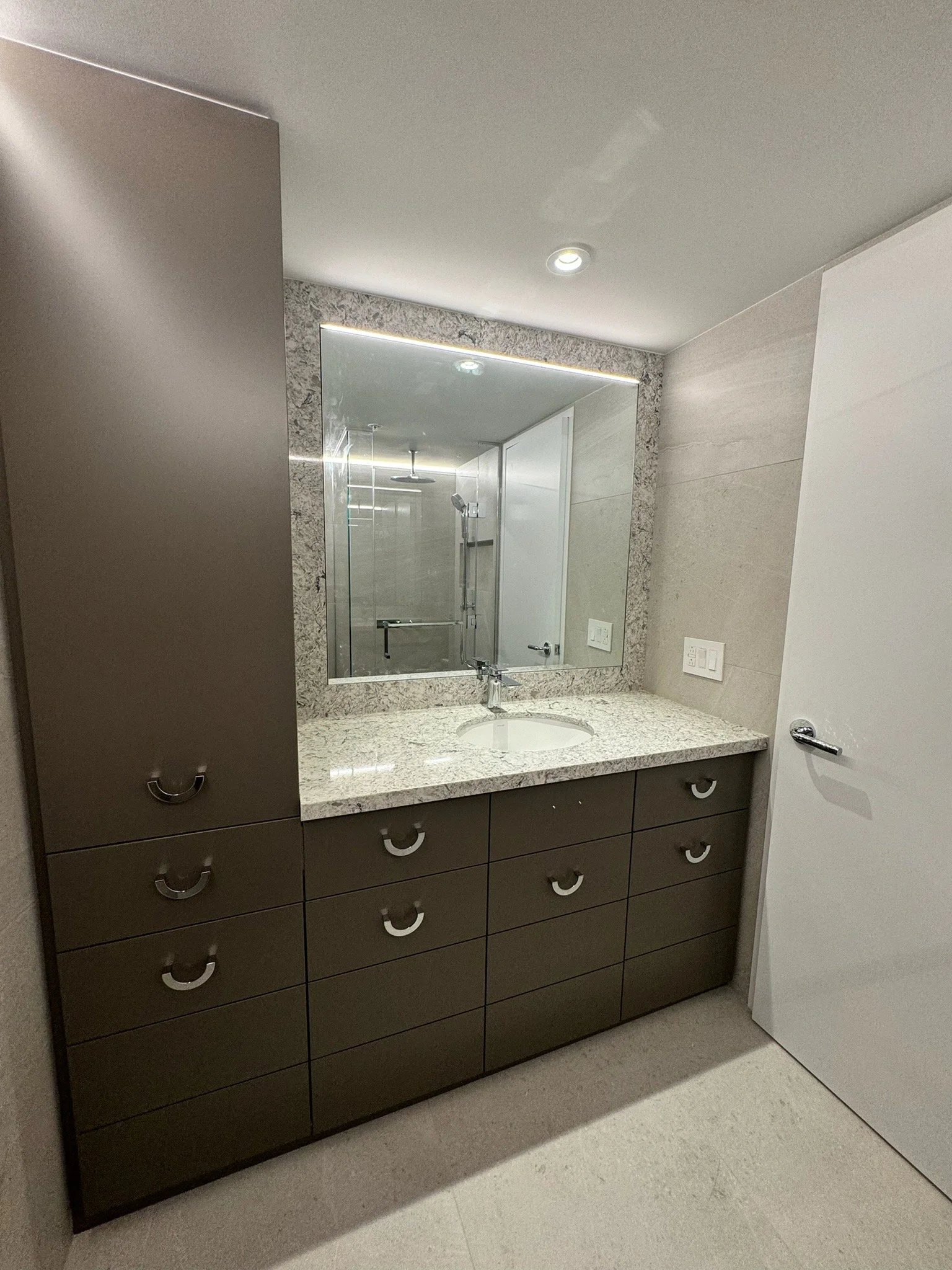 Bathroom vanity with dark brown cabinets, a white granite countertop, a round under-mount sink, and a large mirror. A faucet is installed above the sink. The wall behind has a speckled gray and white pattern, with light switches and electrical outlet