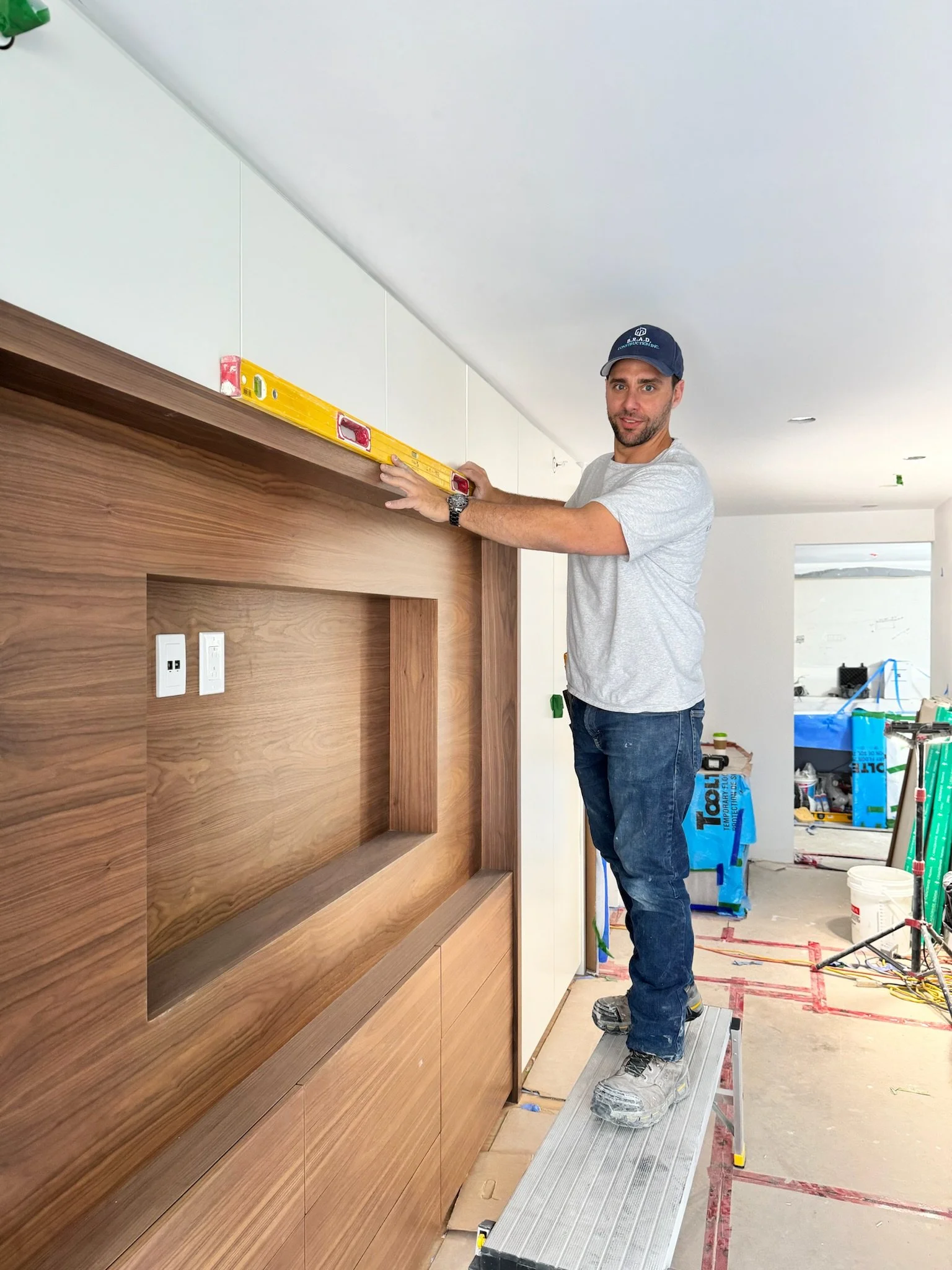A man standing on a small platform practicing measuring a wooden wall with a level, in an indoor construction or renovation site.