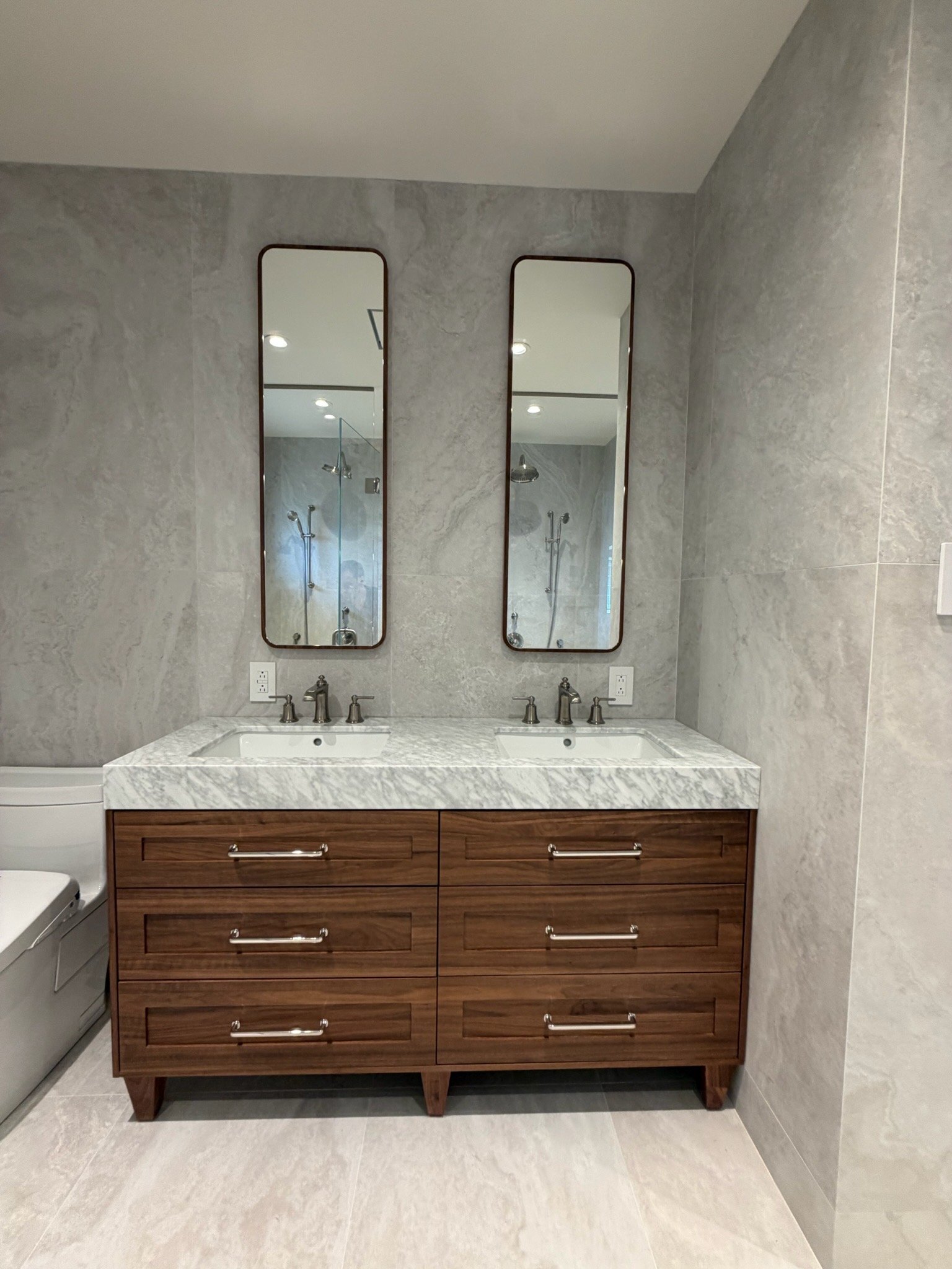 Modern bathroom vanity with a marble countertop, two sinks, and two tall mirrors. The vanity is made of wood with multiple drawers, and the background features light grey marble tiles.