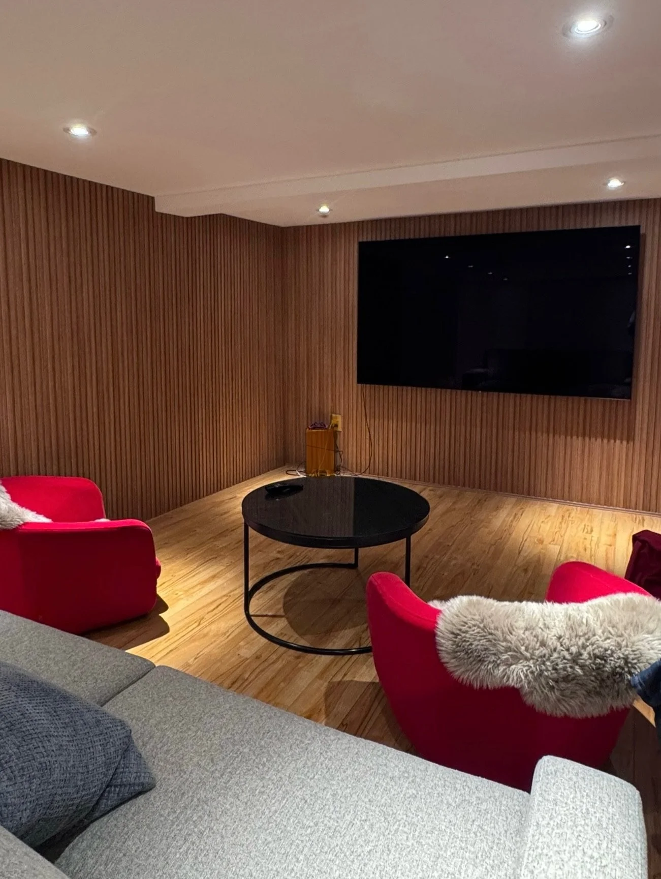 Living room with a big flat-screen TV mounted on a wooden wall, red armchairs with fur throws, a gray sofa with pillows, a black round coffee table, wood flooring, and ceiling lights.