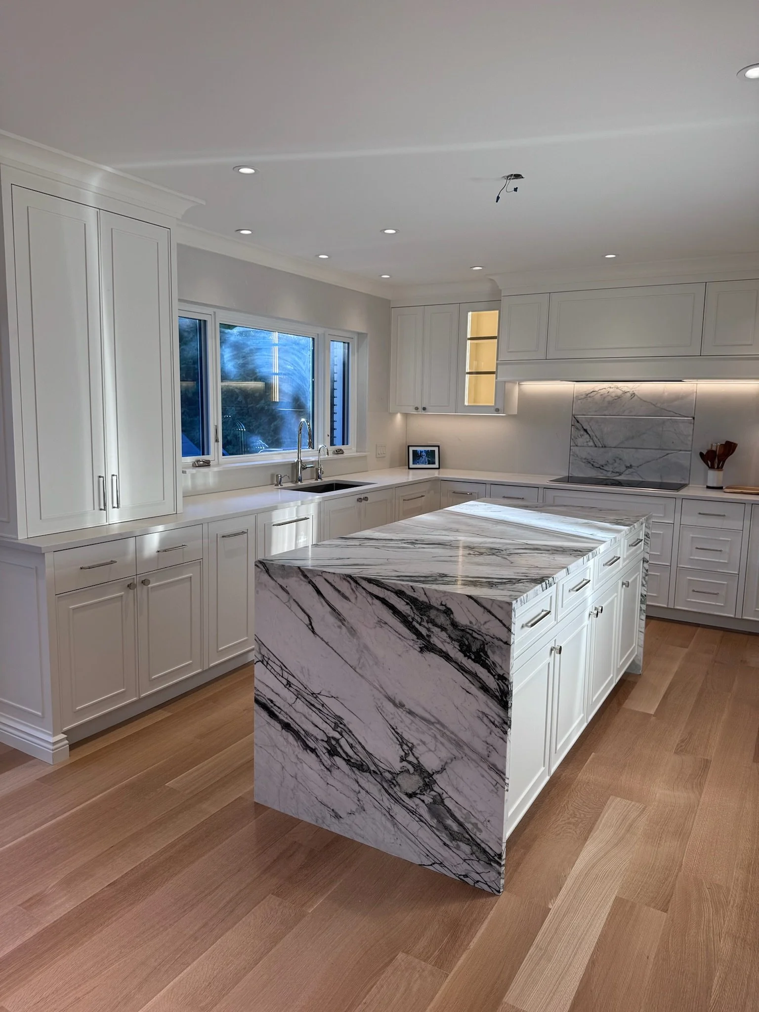 Modern kitchen with white cabinets, marble island, large window, and hardwood floors.