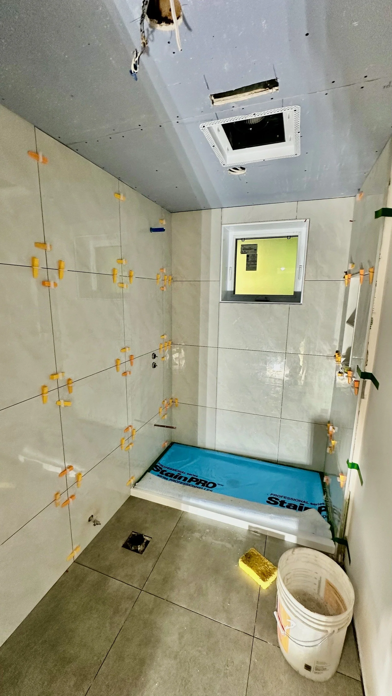 A bathroom under renovation with tiled walls, some tiles are being installed with spacers, a small window, and construction tools and materials on the floor.