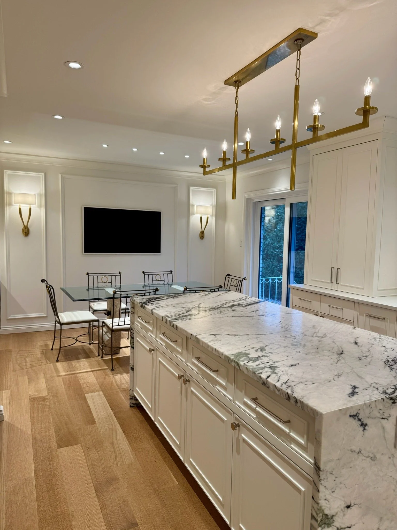 A modern kitchen with a marble countertop island, white cabinetry, gold chandelier, and a dining area with metal chairs, wall-mounted lamps, and a sliding glass door leading outside.
