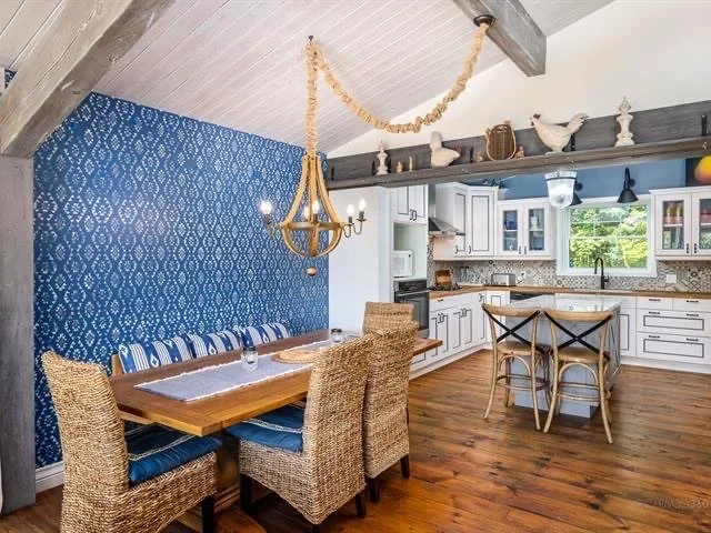 Dining area with a wooden table, wicker chairs, navy and white striped cushions, a blue accent wall with a floral pattern, and a rustic chandelier. Kitchen in the background with white cabinets, a window, and wooden countertops.