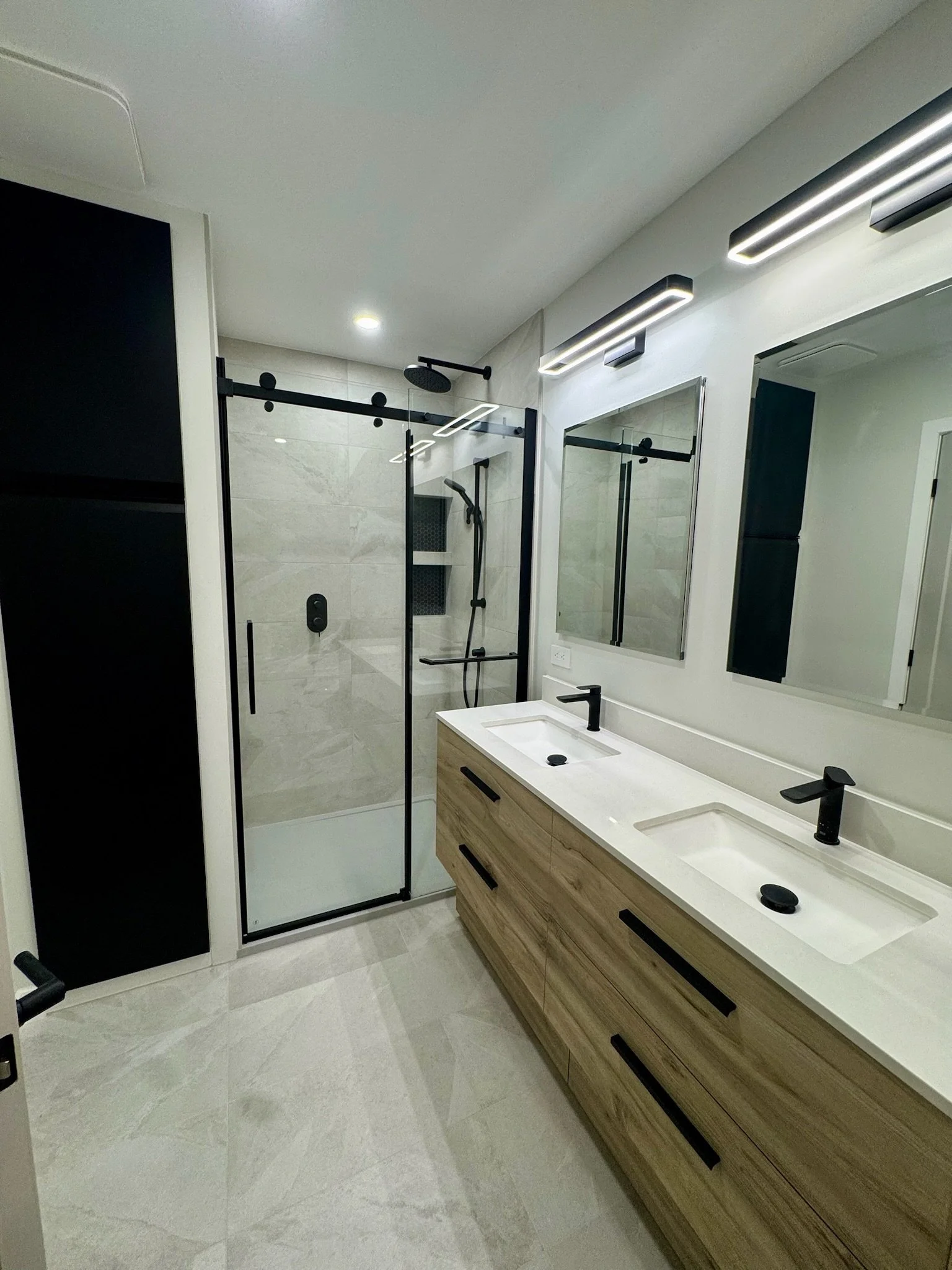 Modern bathroom with double sinks, large mirror, light wood vanity, glass-enclosed shower with black fixtures, and bright overhead lighting.