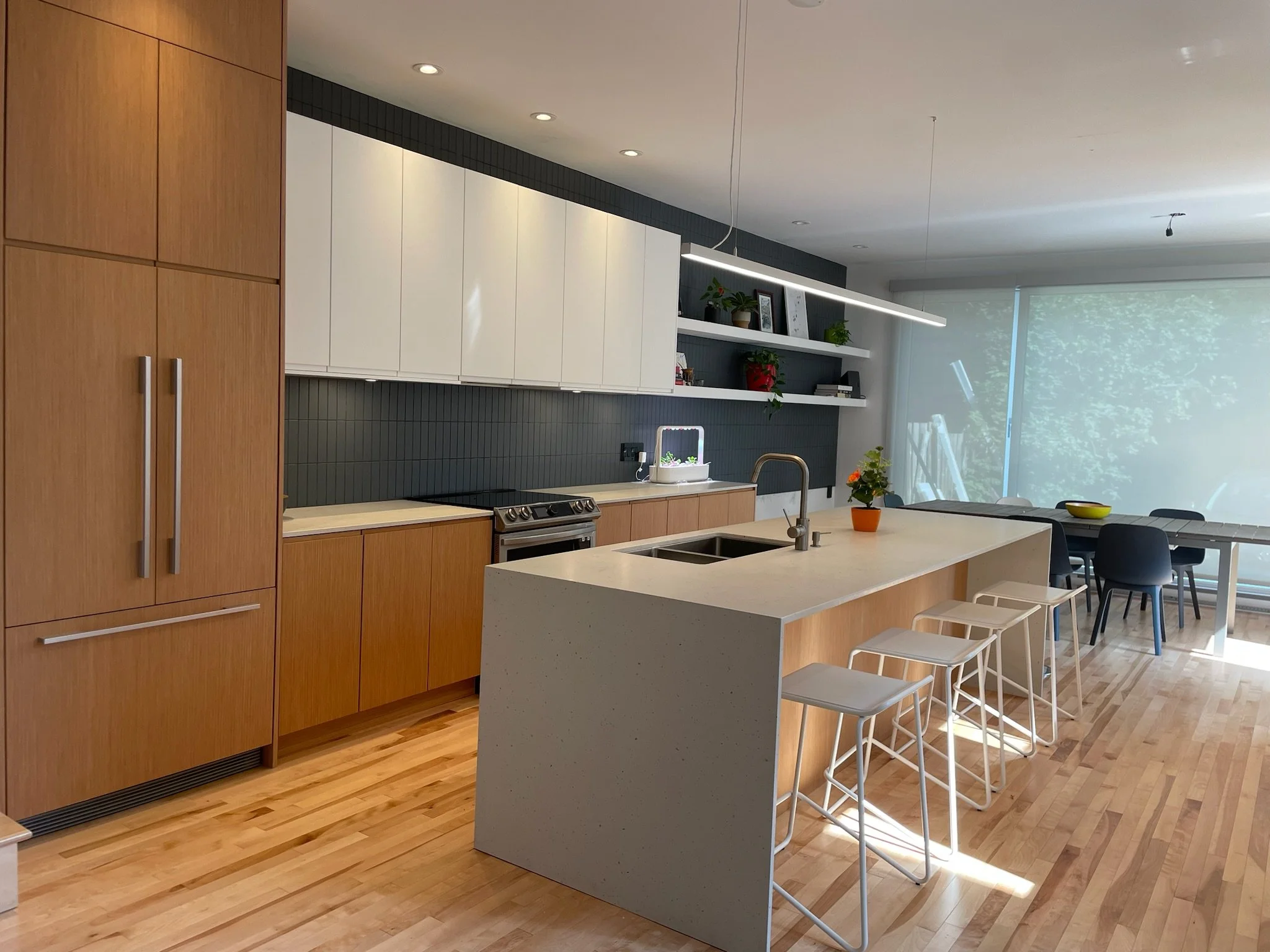 Modern kitchen with wooden cabinets, a white kitchen island with a built-in sink, and open shelves with plants and decor. A dining area with black chairs and large windows with blinds are in the background.