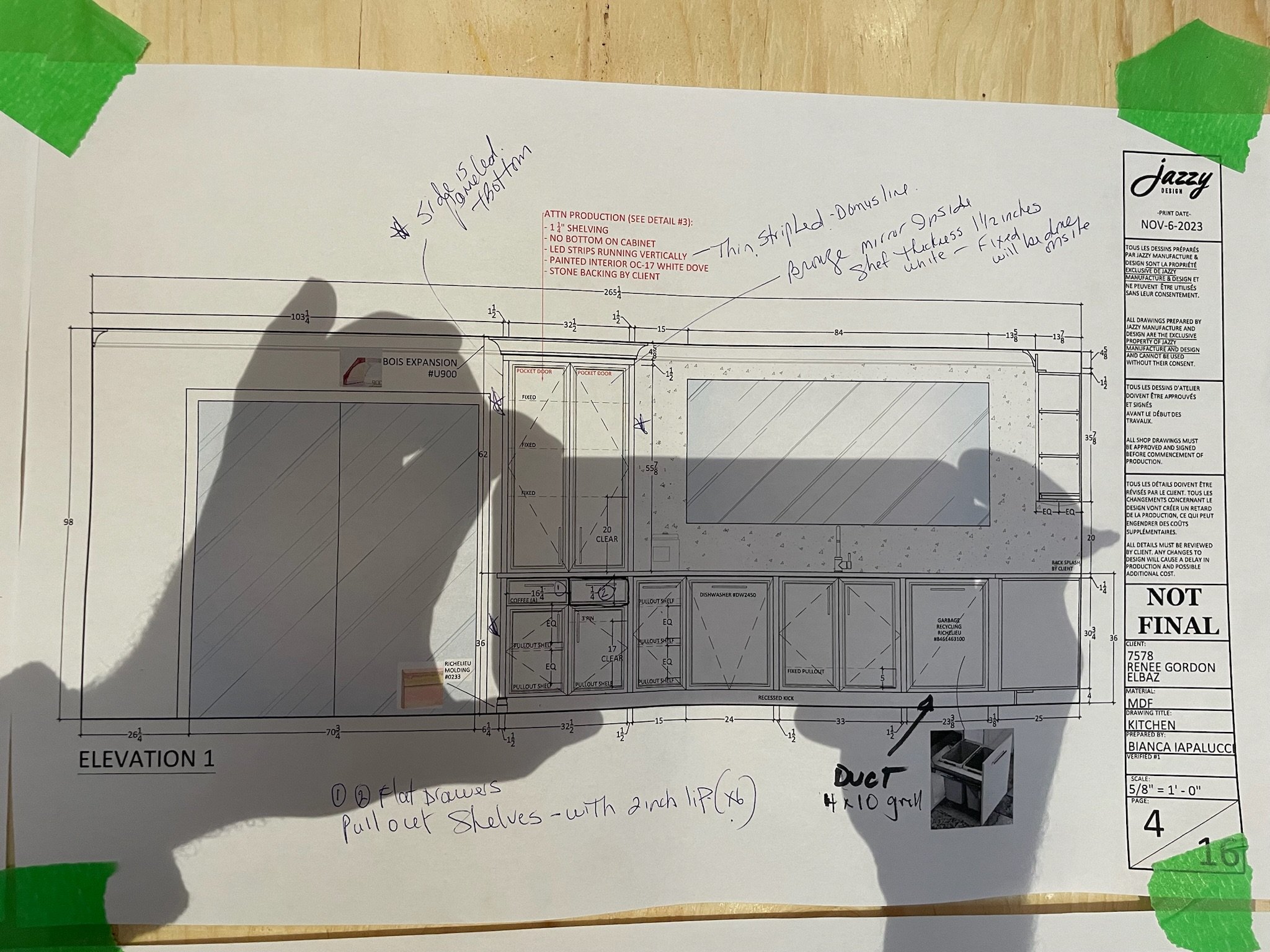 Architectural blueprint of a kitchen layout with measurements, notes, and annotations, taped at green corners to a wooden surface.