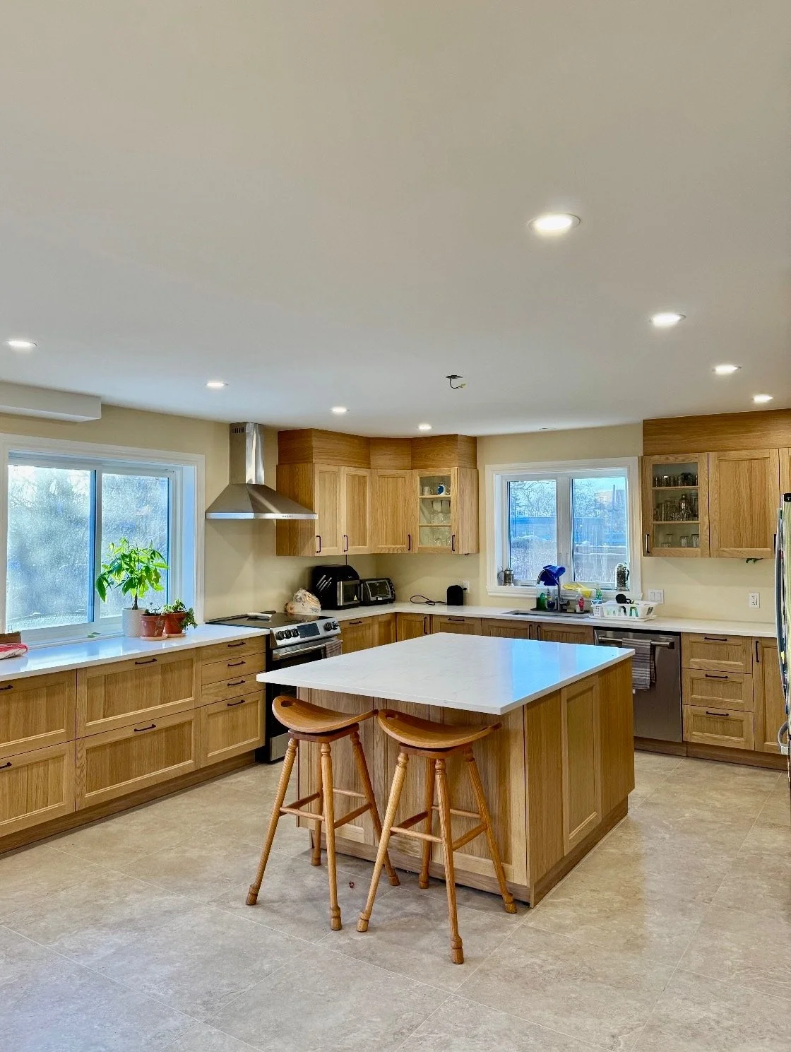 A spacious kitchen with wooden cabinets, a central island, two bar stools, and large windows letting in natural light.
