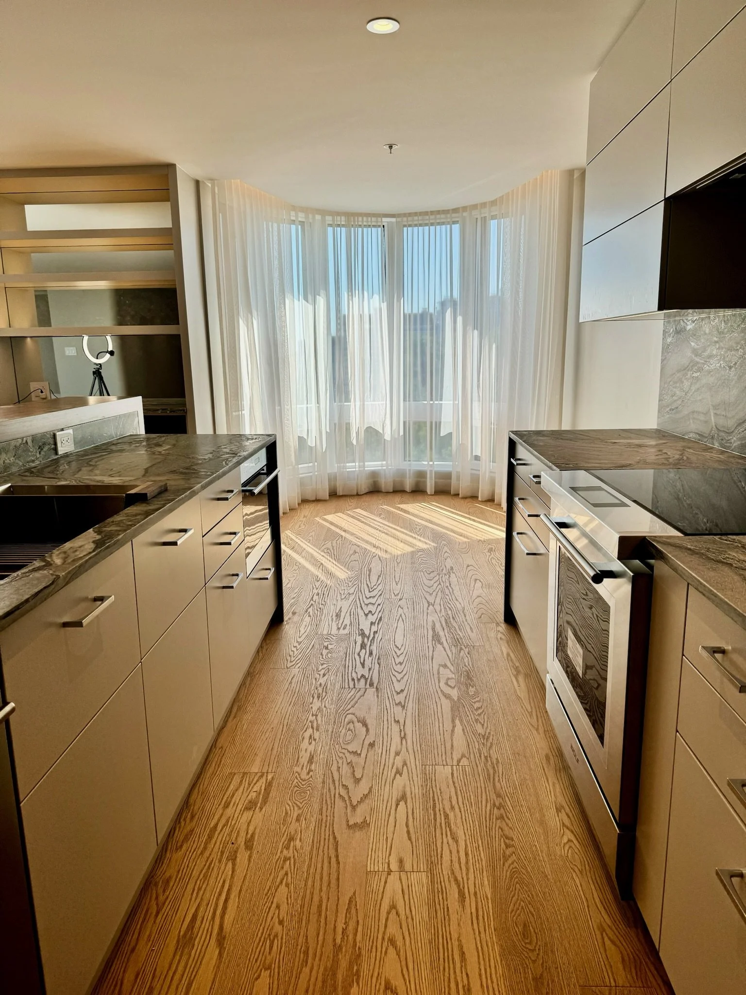 Modern kitchen with wooden flooring, beige cabinets, marble countertops, and large window with white curtains.
