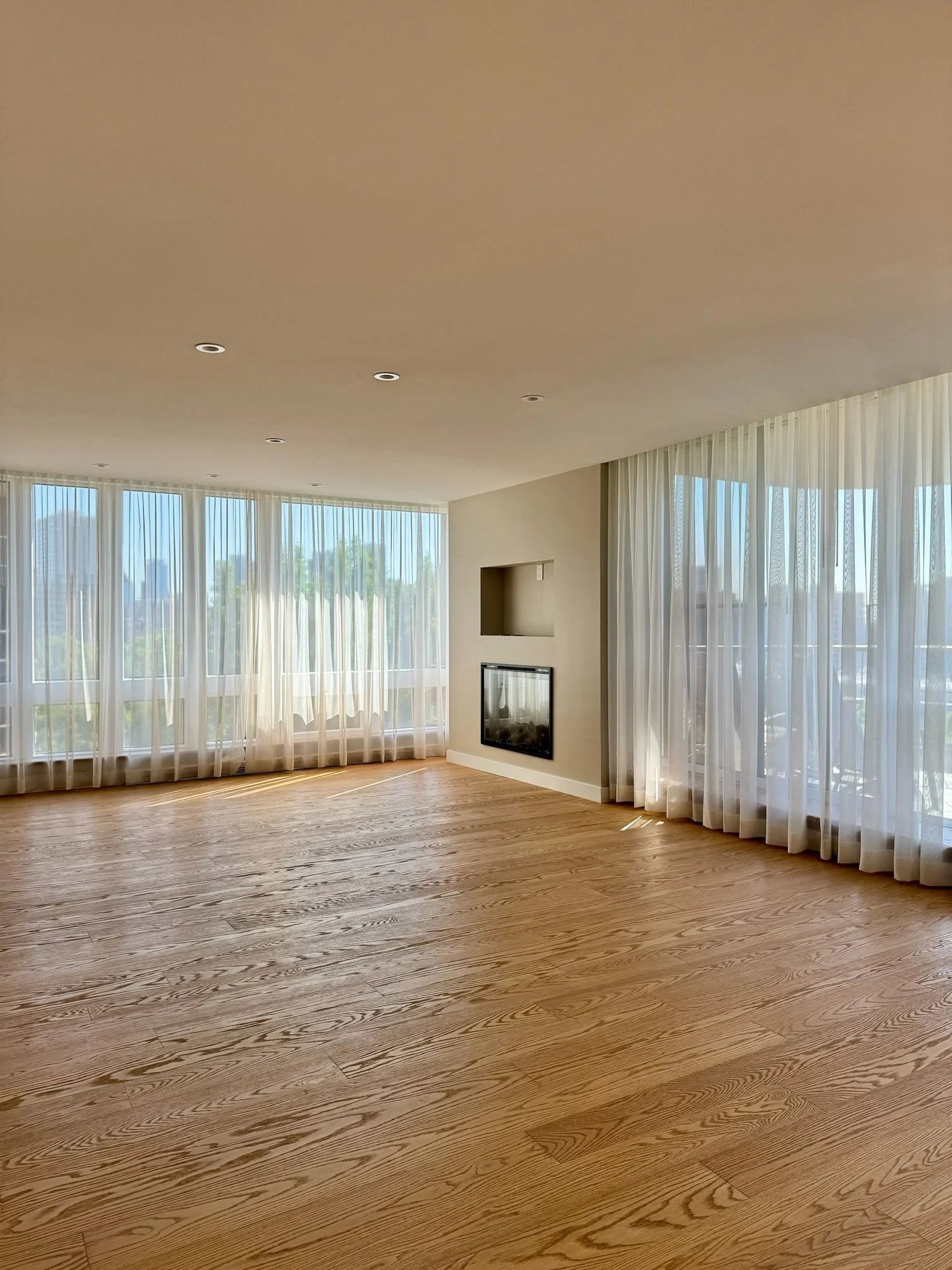 A spacious, modern living room with large windows and white curtains, hardwood floors, a wall-mounted fireplace, and a view of trees and buildings outside.