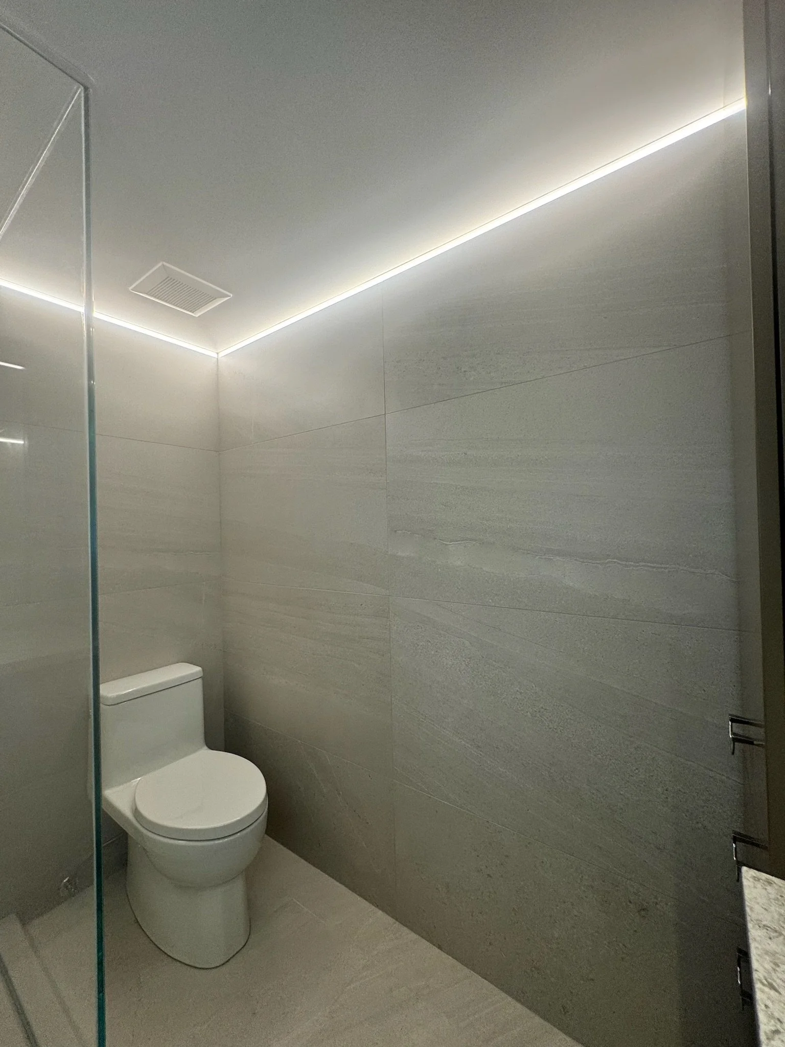 A modern bathroom with light gray tiled walls, a white toilet, a mirror on the left, and a glass shower door on the left side.