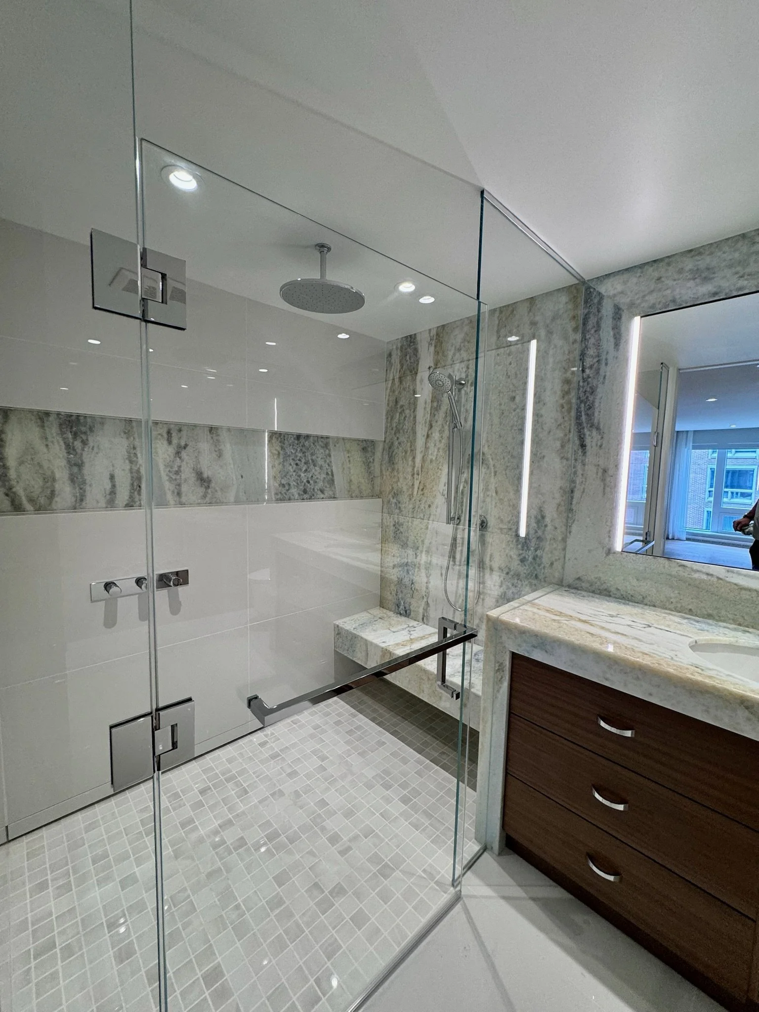 Modern bathroom with marble walls, glass shower enclosure, rainfall shower head, handheld shower, window, and wooden vanity with three drawers.