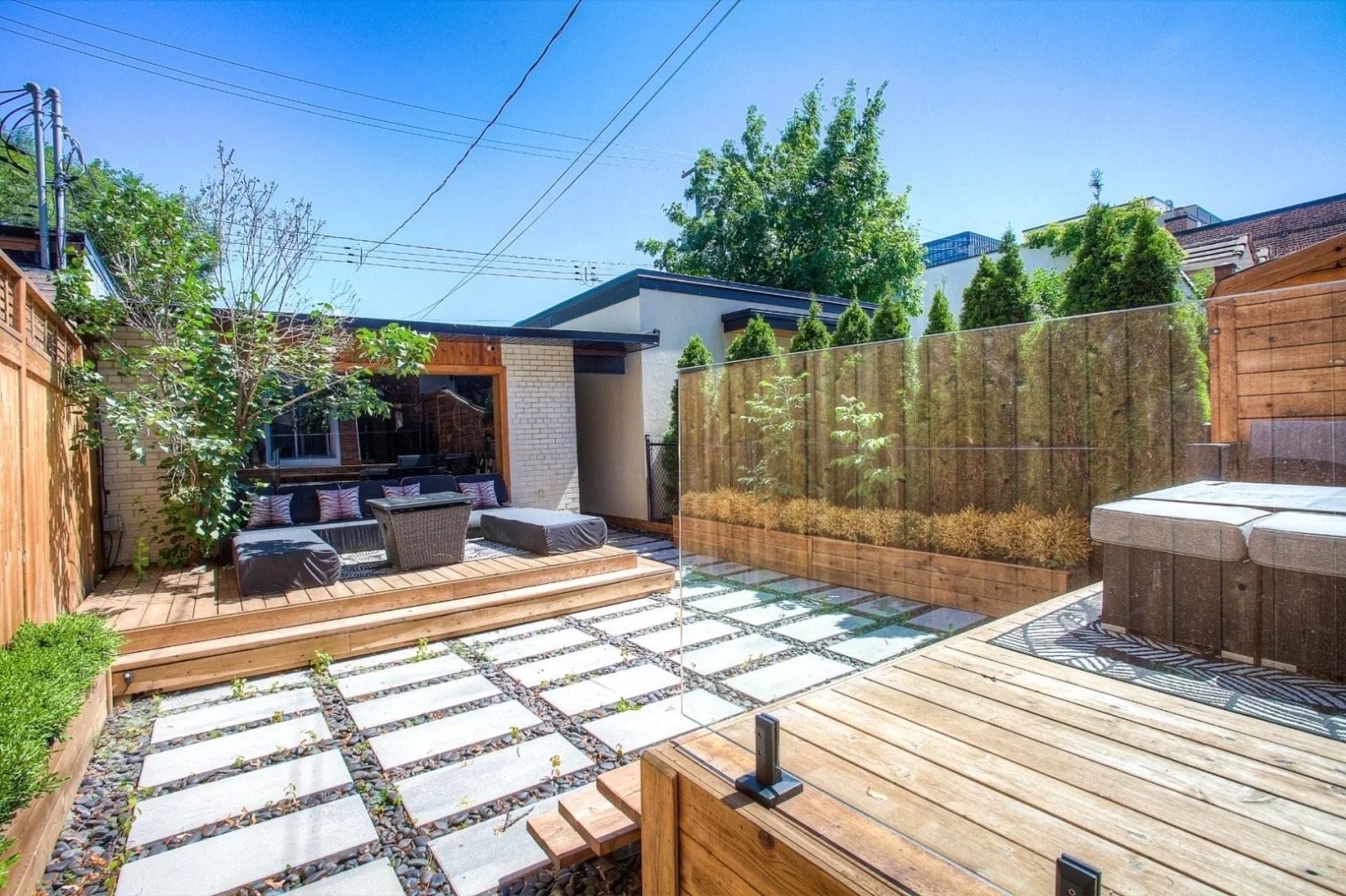 Modern backyard patio with wooden decking, outdoor seating, green plants, small trees, and a privacy fence under a clear blue sky.