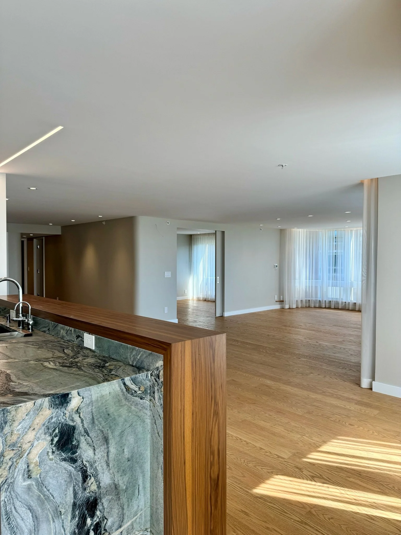 Modern open-concept living space with hardwood floors, large windows with sheer curtains, a kitchen island with a marble and wood finish, and white walls.