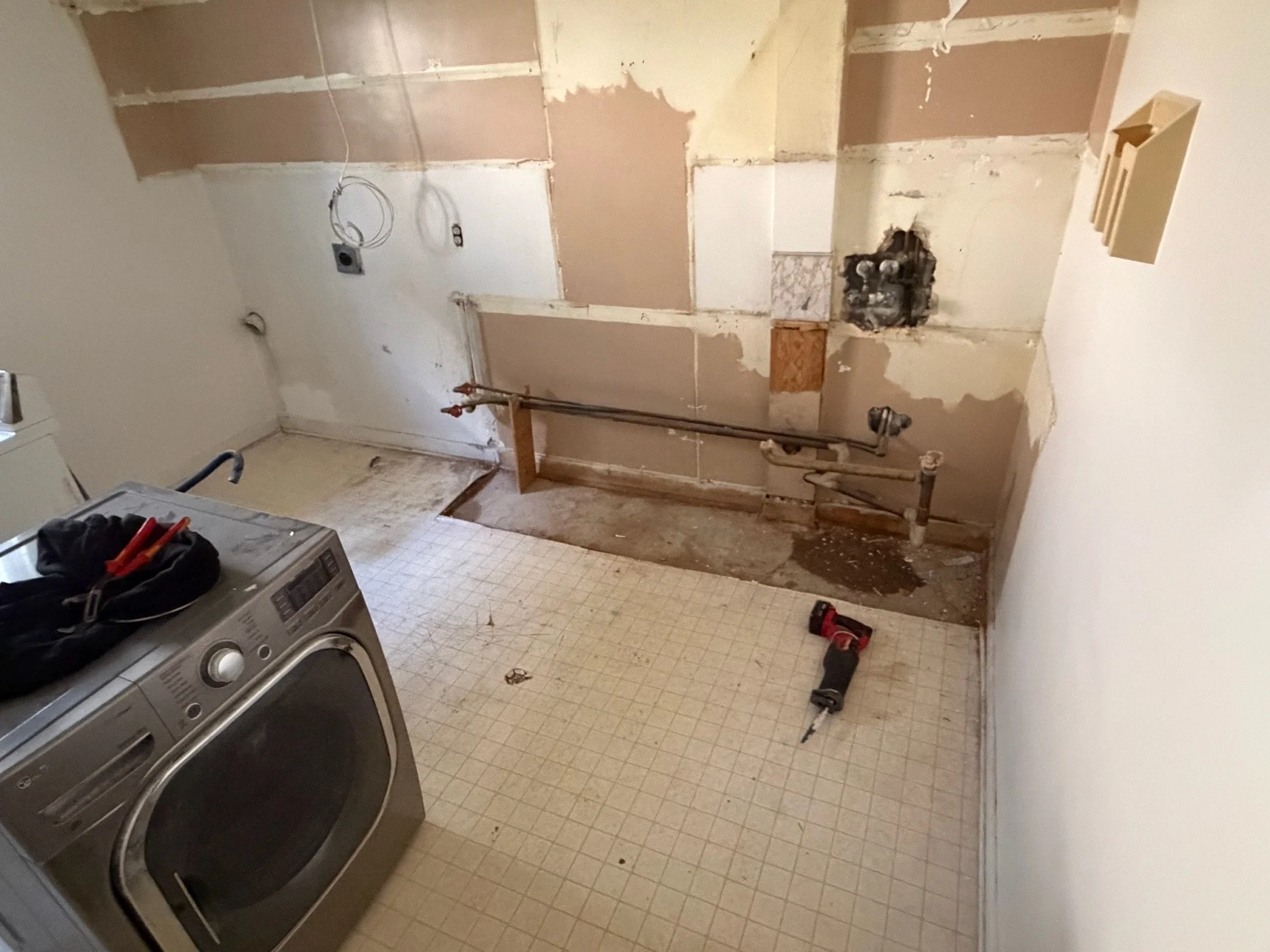 Laundry room with removed cabinets and drywall, exposed plumbing, a washing machine with tools on top, and a floor with a small section of removed tiles.