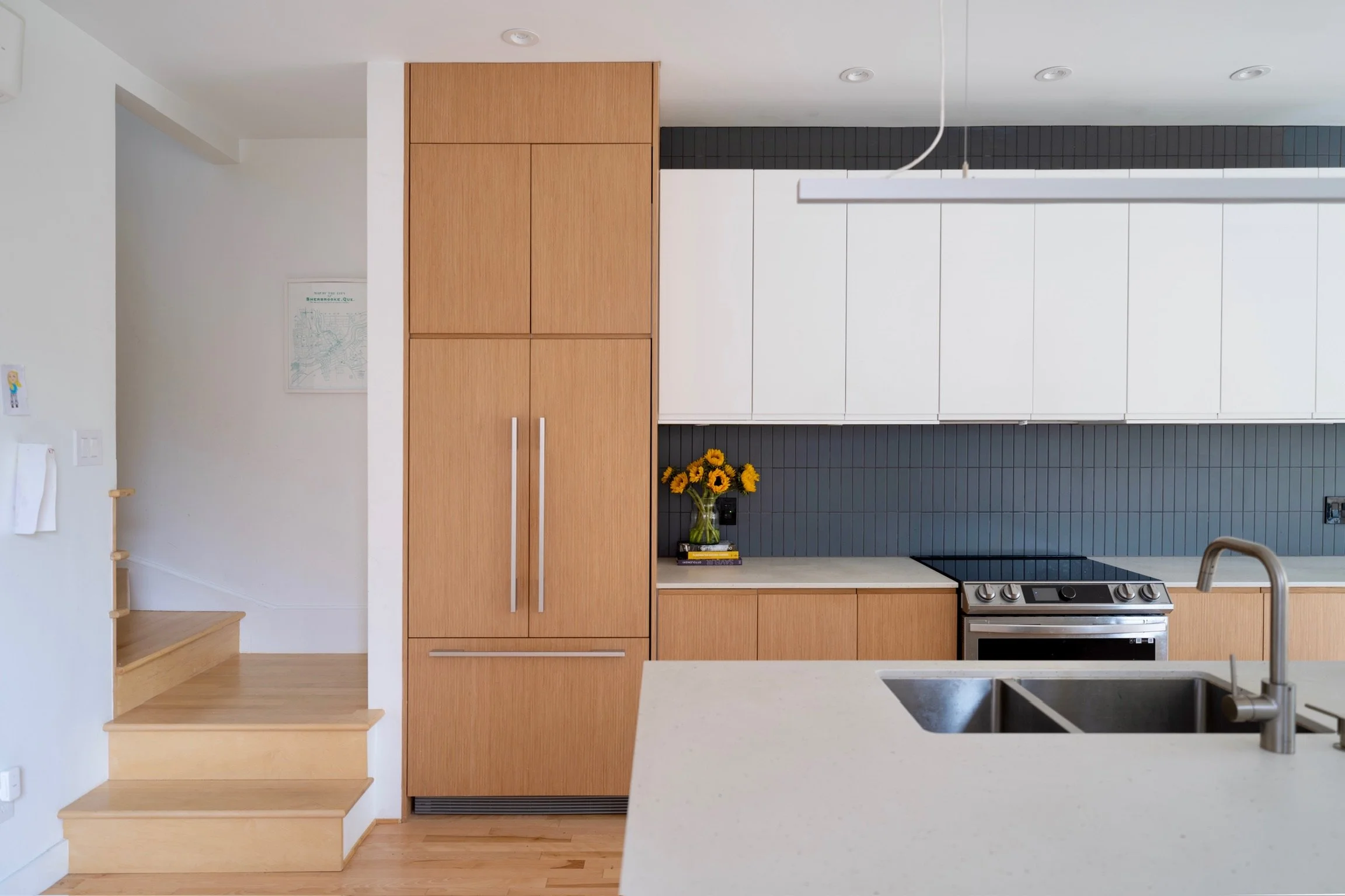 A modern kitchen with white cabinets, a grey tiled backsplash, a stainless steel oven, a black cooktop, a white countertop, a stainless steel sink, a vase with sunflowers, and a set of wooden stairs.