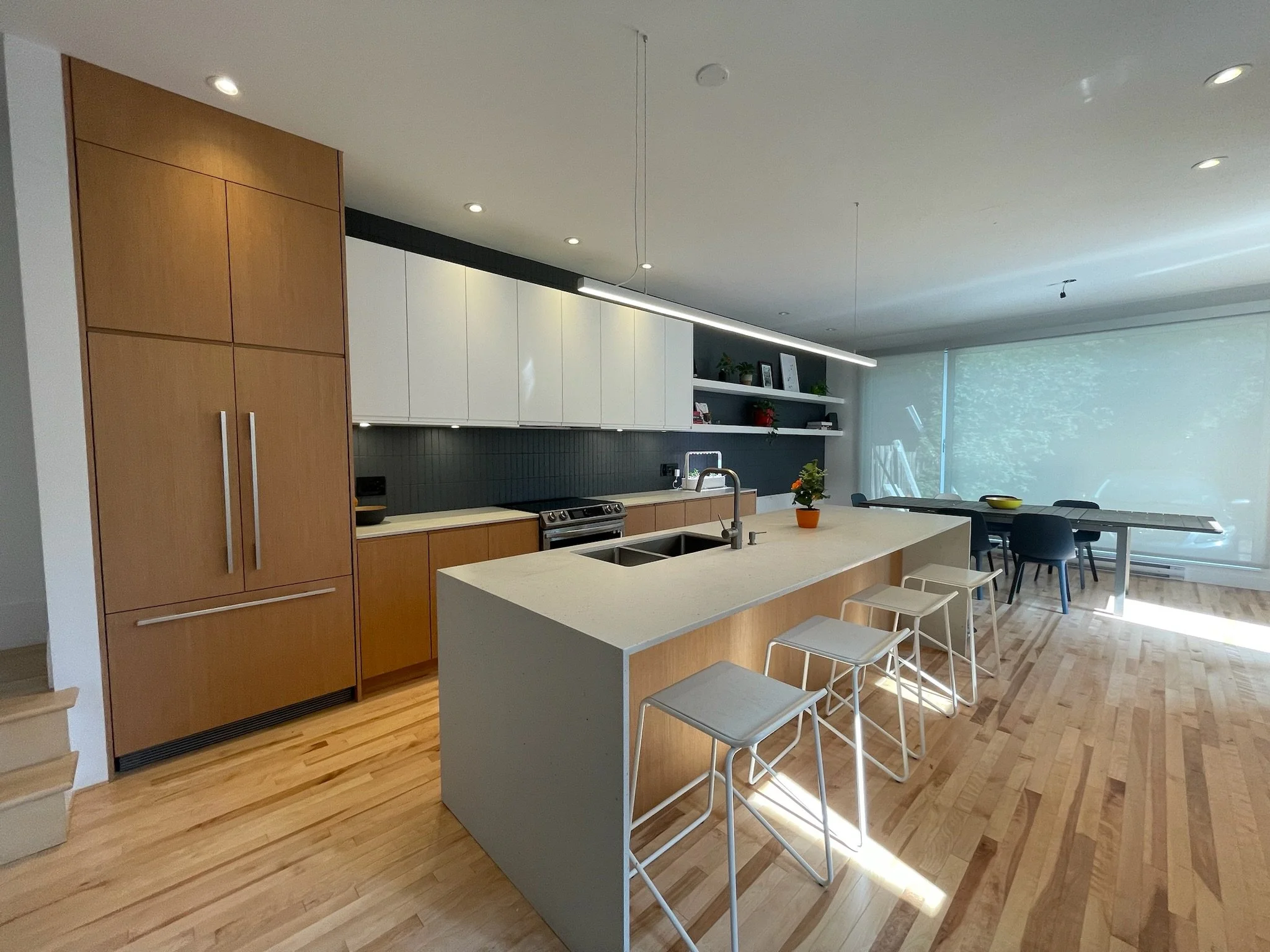 Modern kitchen with wooden cabinets, a light-colored island with bar stools, and a large window with a view of greenery outside.