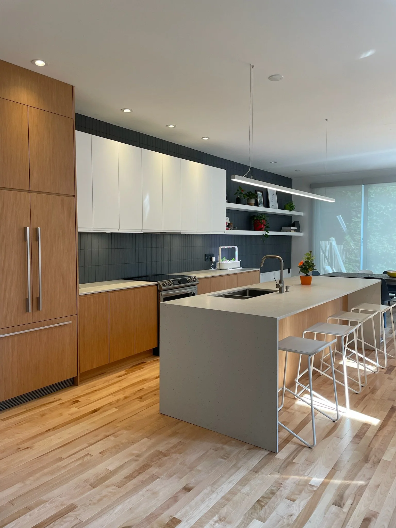Modern kitchen with wooden cabinets, a white kitchen island, and open shelves with decorative items and plants. Large windows in the background let in natural light.