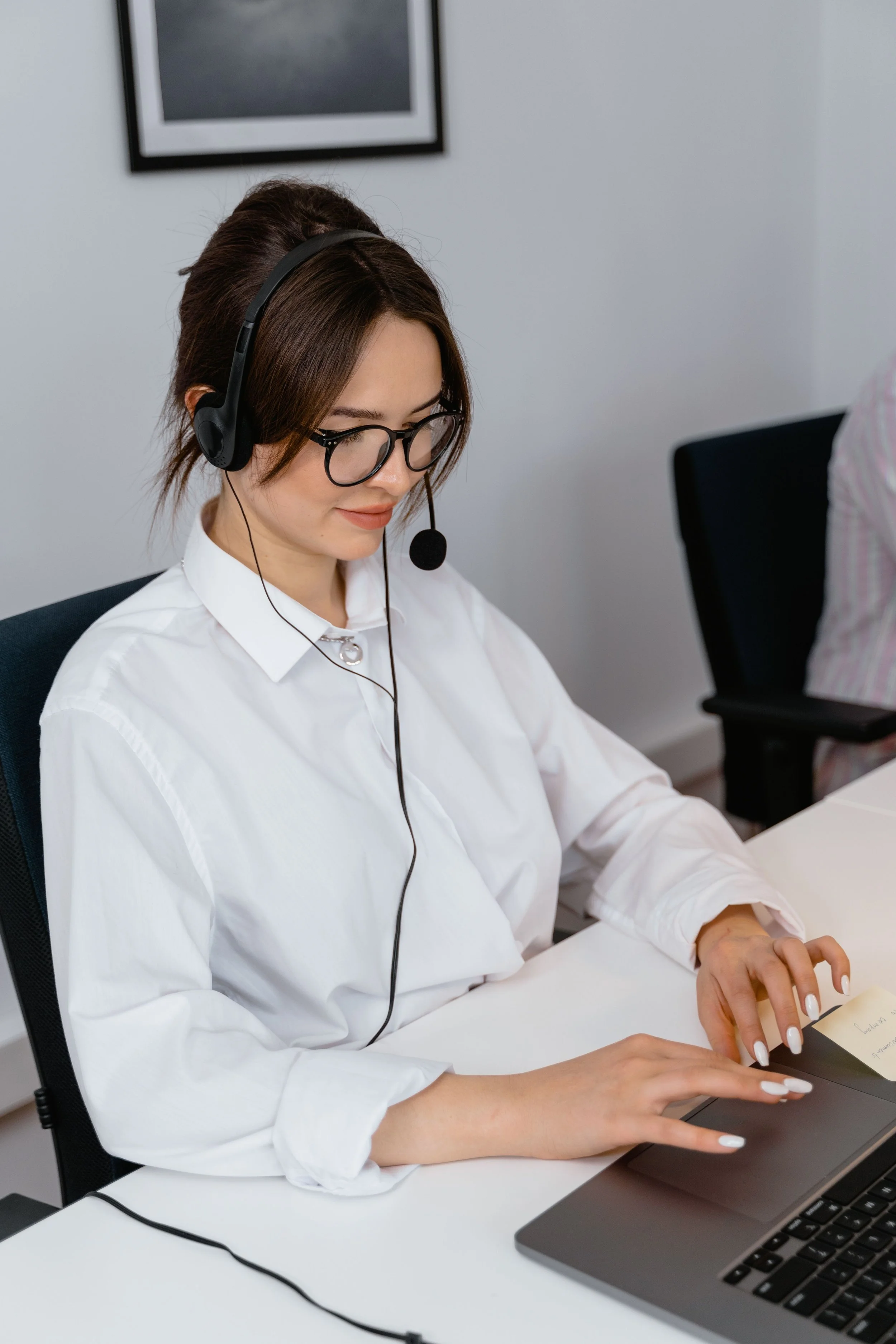 A woman wearing glasses and a white shirt using a laptop, with a headset on, sitting at a desk in an office.