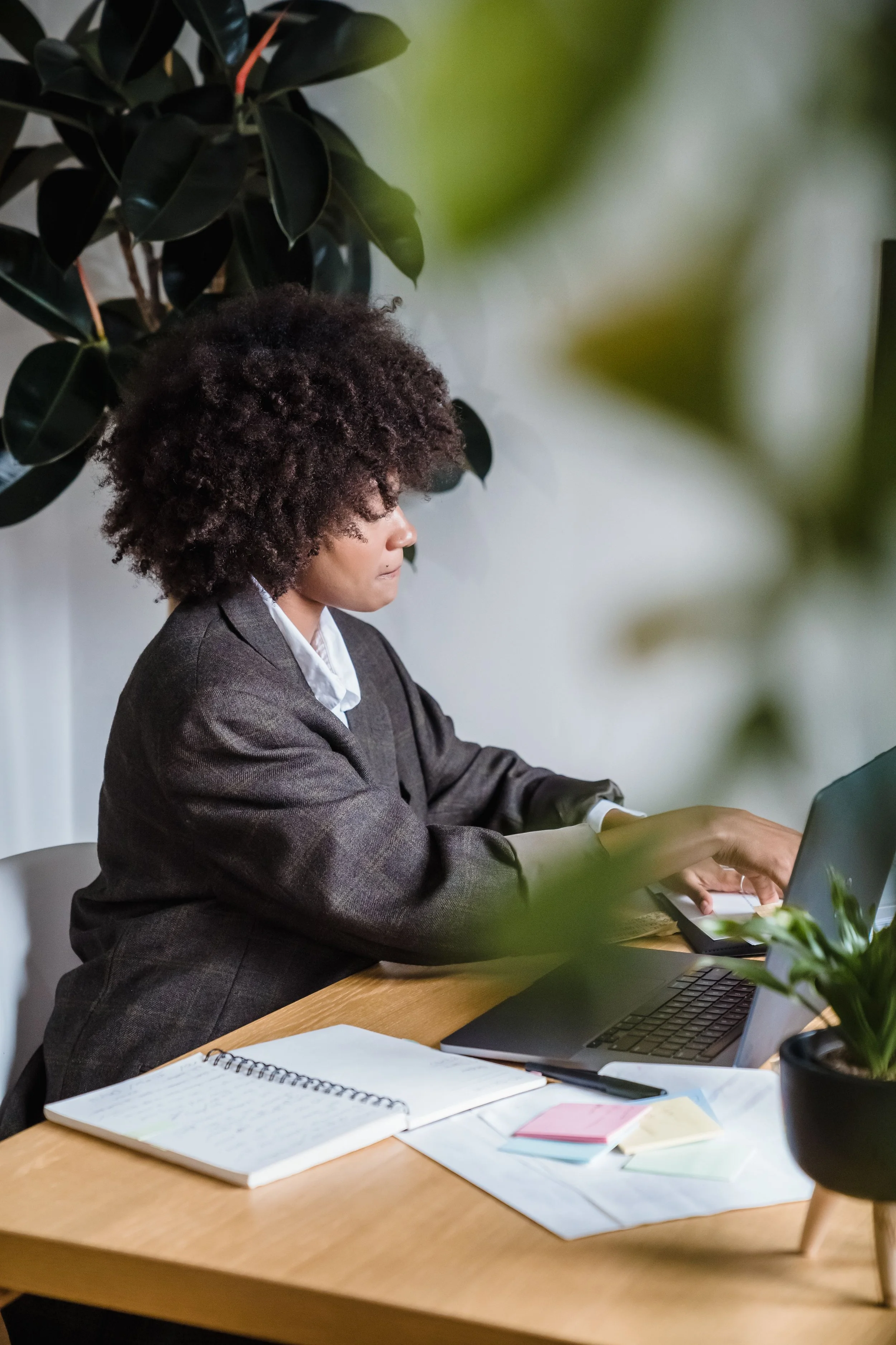 A woman with curly hair wearing a gray suit, sitting at a wooden table, playing a digital piano, with a plant and some notebooks on the table.