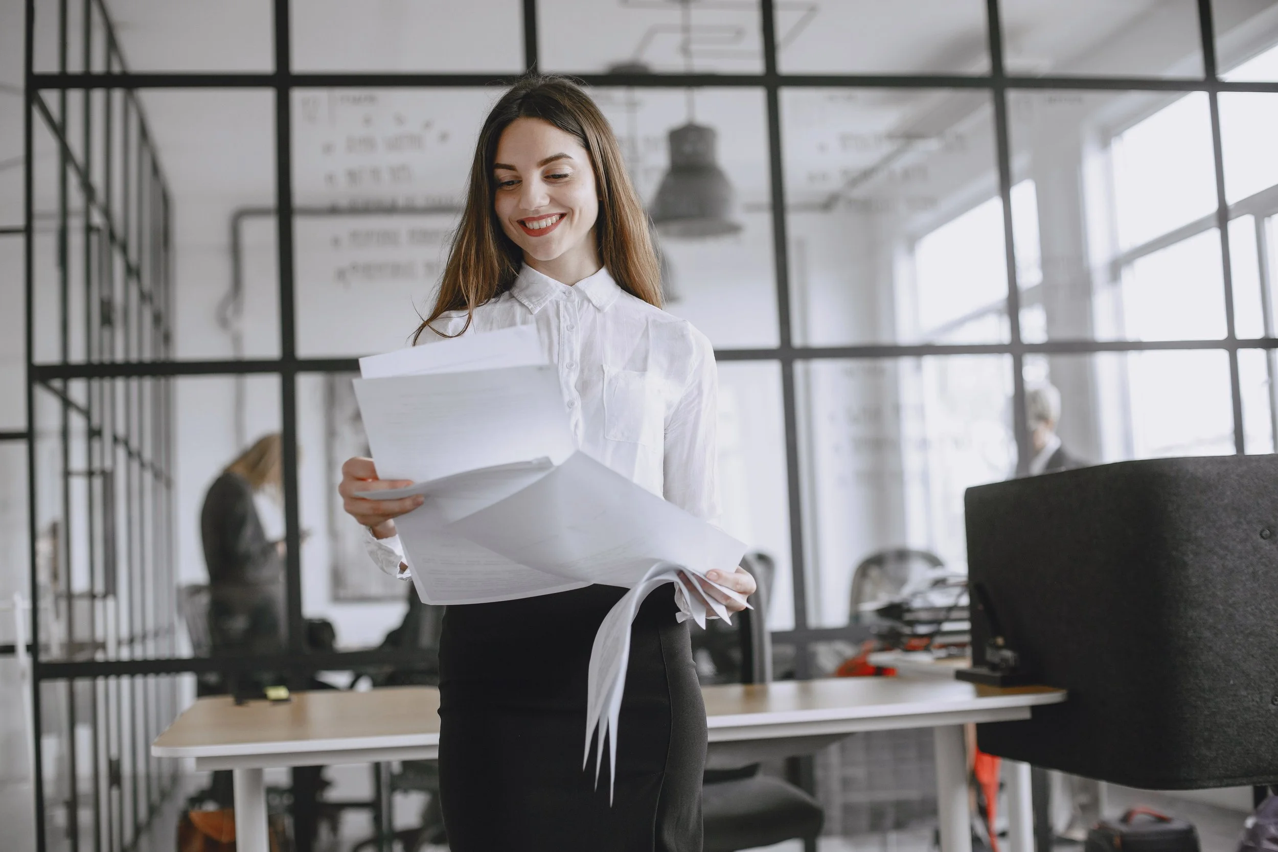 A woman with long brown hair and fair skin, wearing a white blouse, is smiling while holding a stack of papers in a modern office space with large windows and black metal framing.