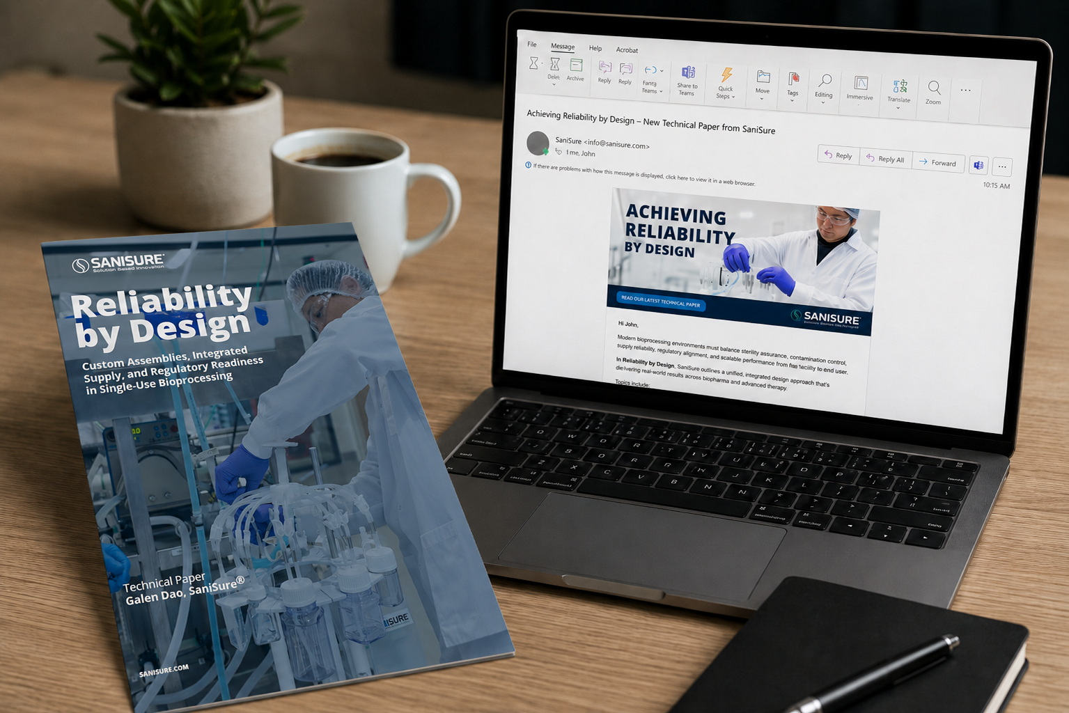 A printed technical paper titled 'Reliability by Design' showing a scientist working in a laboratory, placed on a desk next to a laptop displaying an email with the same paper overview, a coffee mug, a potted plant, and a black notebook with a pen.