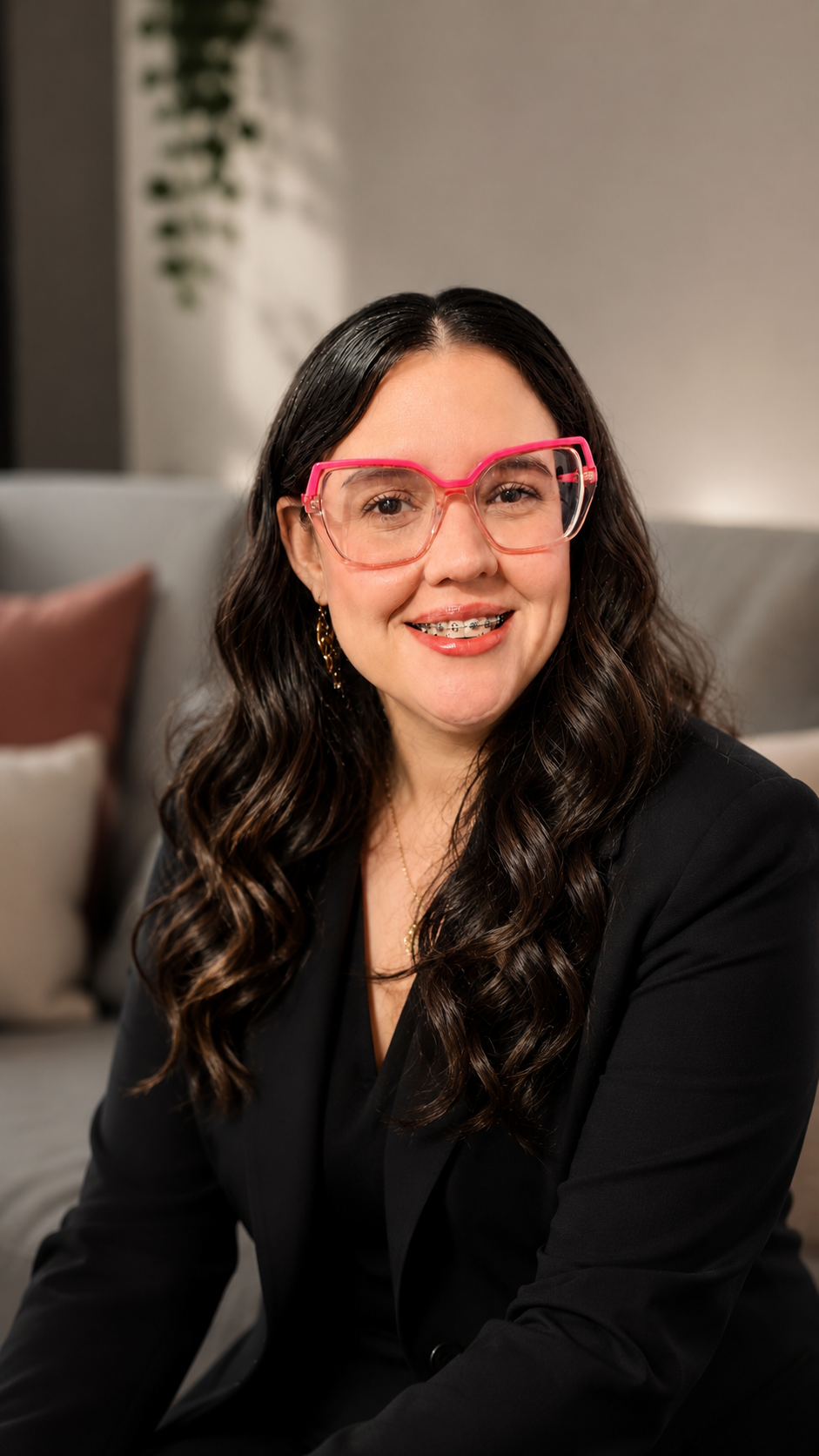 A woman with long dark wavy hair wearing pink glasses, braces, a black blazer, gold earrings, and a necklace, sitting on a sofa in a room with pillows and a blurred background.