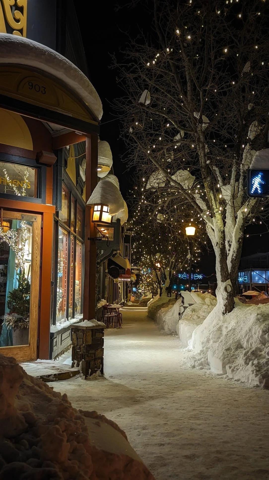 A snowy sidewalk at night with decorated trees and illuminated storefronts. Winter Wonderland. Denver to Ski Resorts. Steamboat Springs. Ski Transportation. Airport Hotel Transfers.