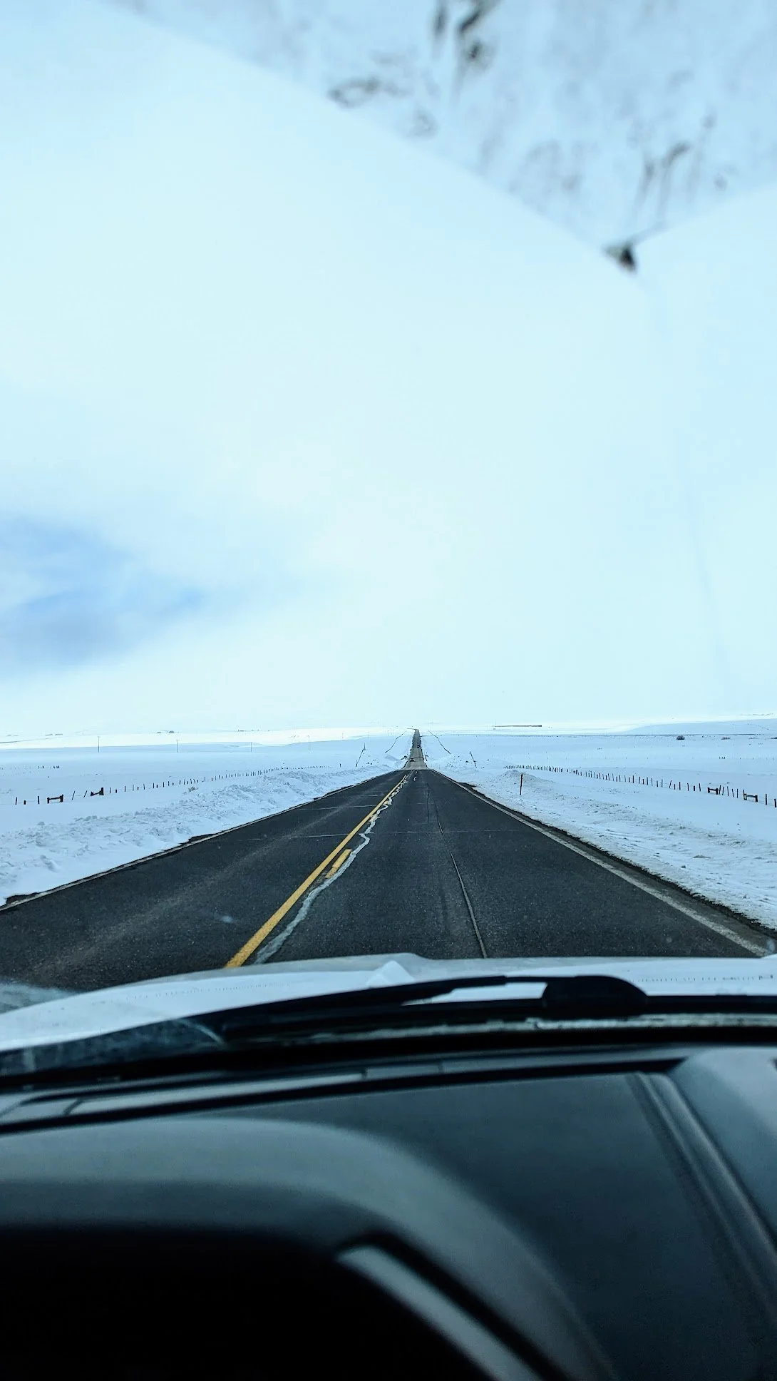 View from inside a vehicle looking at a long, straight, snow-covered road in a rural area with snow on both sides, under a cloudy sky. Colorado. Steamboat Springs. Mountain Roads. Denver to Steamboat. Denver to Ski Mountains.