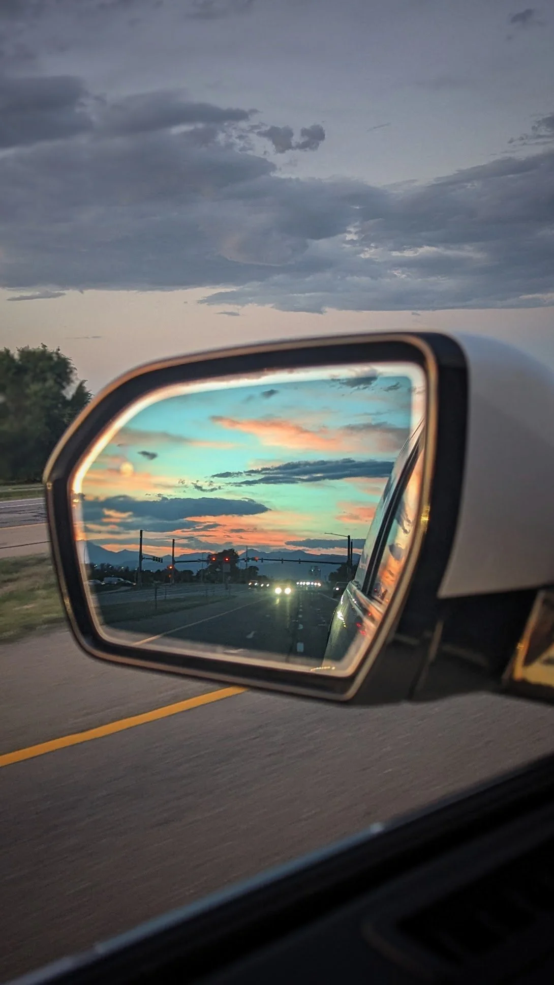 View of a sunset sky with colorful clouds reflected in a car's side mirror as cars drive down a highway. Comfortable rides. Airport Transfers from Boulder County to DIA