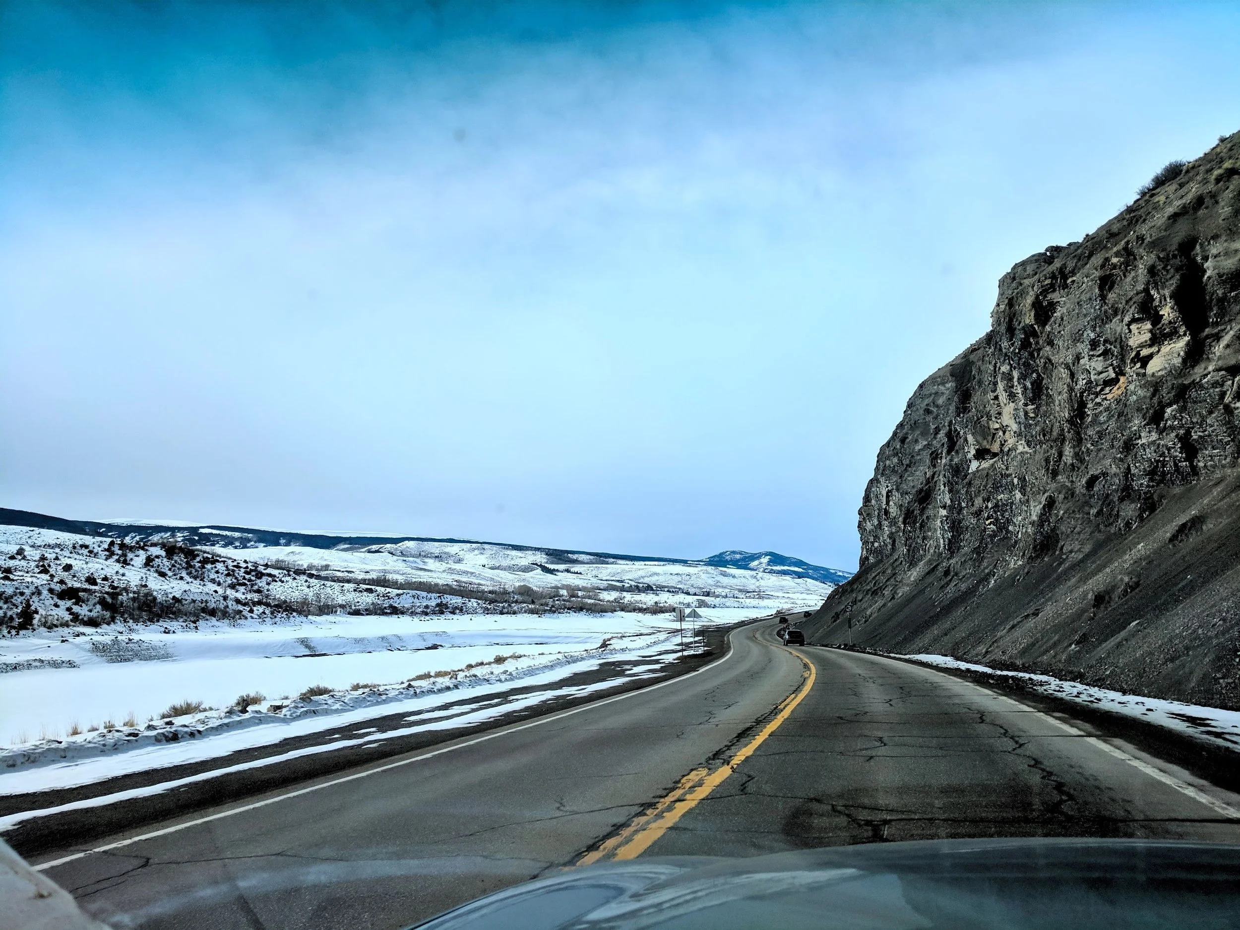 A winding mountain road with snow on the ground and a car driving on it, next to a rocky hillside under a cloudy sky. Destination Steamboat Springs.